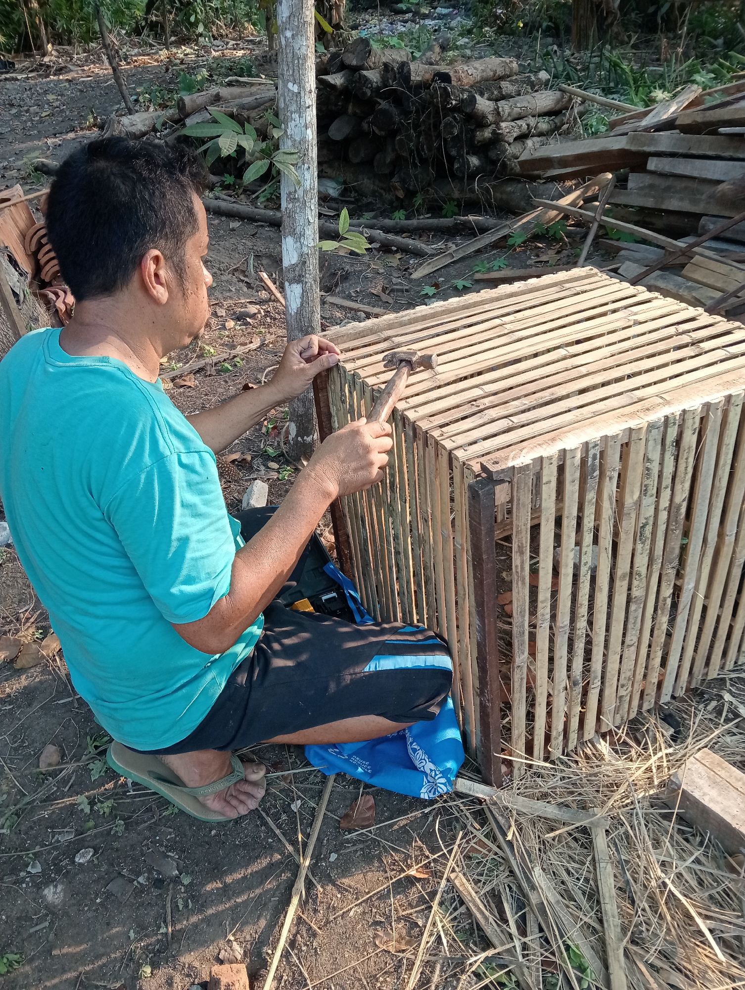 In Muara, Indonesia, a man is shown building a cage outdoors. He is kneeling on the ground, wearing a turquoise t-shirt and dark shorts. His hands are occupied with a hammer and what appears to be a nail, as he works on the bamboo structure of the cage. The cage itself is made of vertical bamboo poles and has a slatted top. The surrounding environment is natural and somewhat unkempt, with scattered logs, branches, and dry leaves on the ground. A tall, thin tree stands to the left of the man. The lighting suggests it is daytime, possibly late afternoon, with sunlight casting shadows. The scene appears to be a simple, outdoor workshop setting where manual labor is being performed.