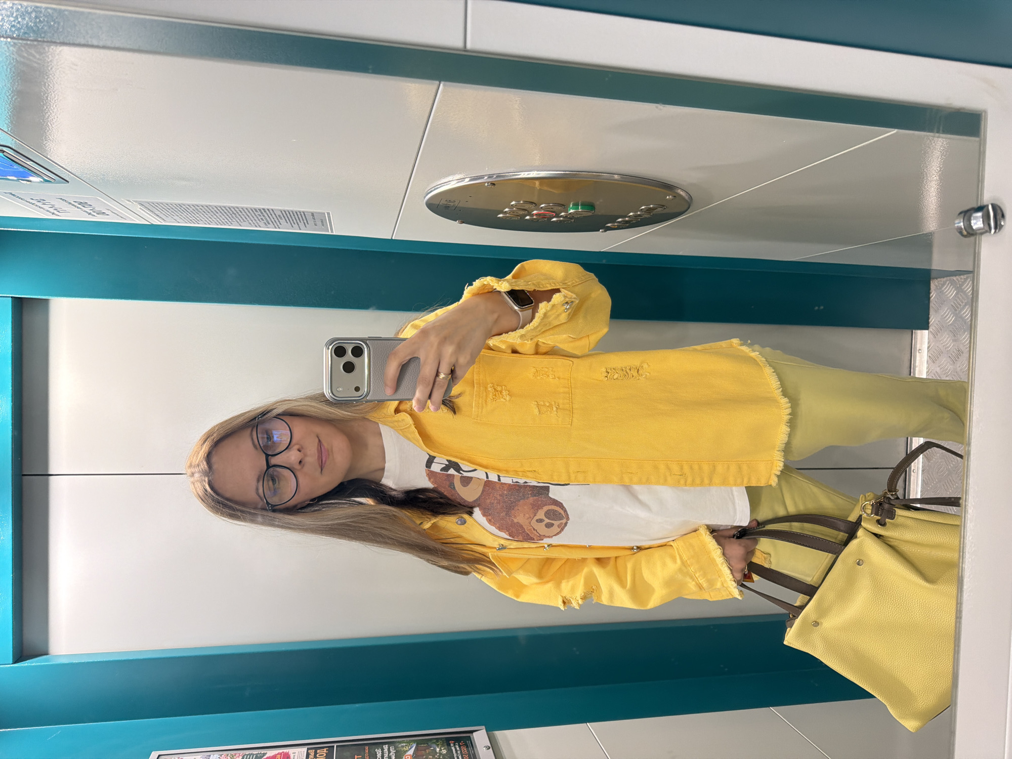 An adult Caucasian female with blonde hair and glasses is standing in an elevator and taking a mirror selfie. She is wearing a yellow denim jacket over a white t-shirt with a graphic print of a teddy bear. Her pants are also yellow. She is carrying a large yellow tote bag with brown handles. The elevator walls are white with teal accents. A metal panel with elevator buttons is visible on the right wall. The floor of the elevator appears to be metal with a diamond plate pattern. The image was taken in an elevator in Izhevsk, Russia.