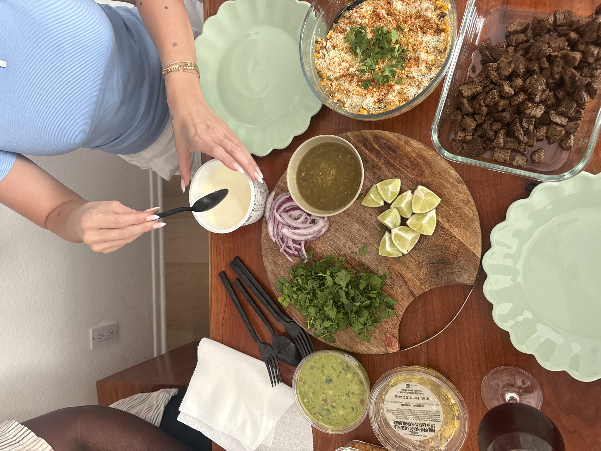A top-down, medium shot depicts a table setting for what appears to be a taco-style meal preparation, possibly in a residential setting in Los Angeles, given the typical house design and plug socket in the lower left of the frame. On a wooden table, there are multiple dishes of food arranged, including a glass baking dish filled with small, dark brown cubes of meat, presumably beef or steak. Adjacent to it is another glass dish containing a dish topped with corn and herbs, with chili powder seasoning. A wooden cutting board serves as a centerpiece, holding a small bowl of green salsa, slices of lime, chopped red onions, and a pile of fresh cilantro. Below this is a container of guacamole and a container of pineapple mango salsa. Two light green ruffled plates are also visible. A fair-skinned person wearing a light blue top and gold bracelet is positioned on the left side of the image. They are holding a black spoon and reaching into a white container, likely sour cream or yogurt. The person is preparing to add the contents of the spoon to the plates. Three black forks are neatly arranged on a white napkin next to the containers. A wine glass filled with a red liquid, possibly wine, is in the lower right corner.