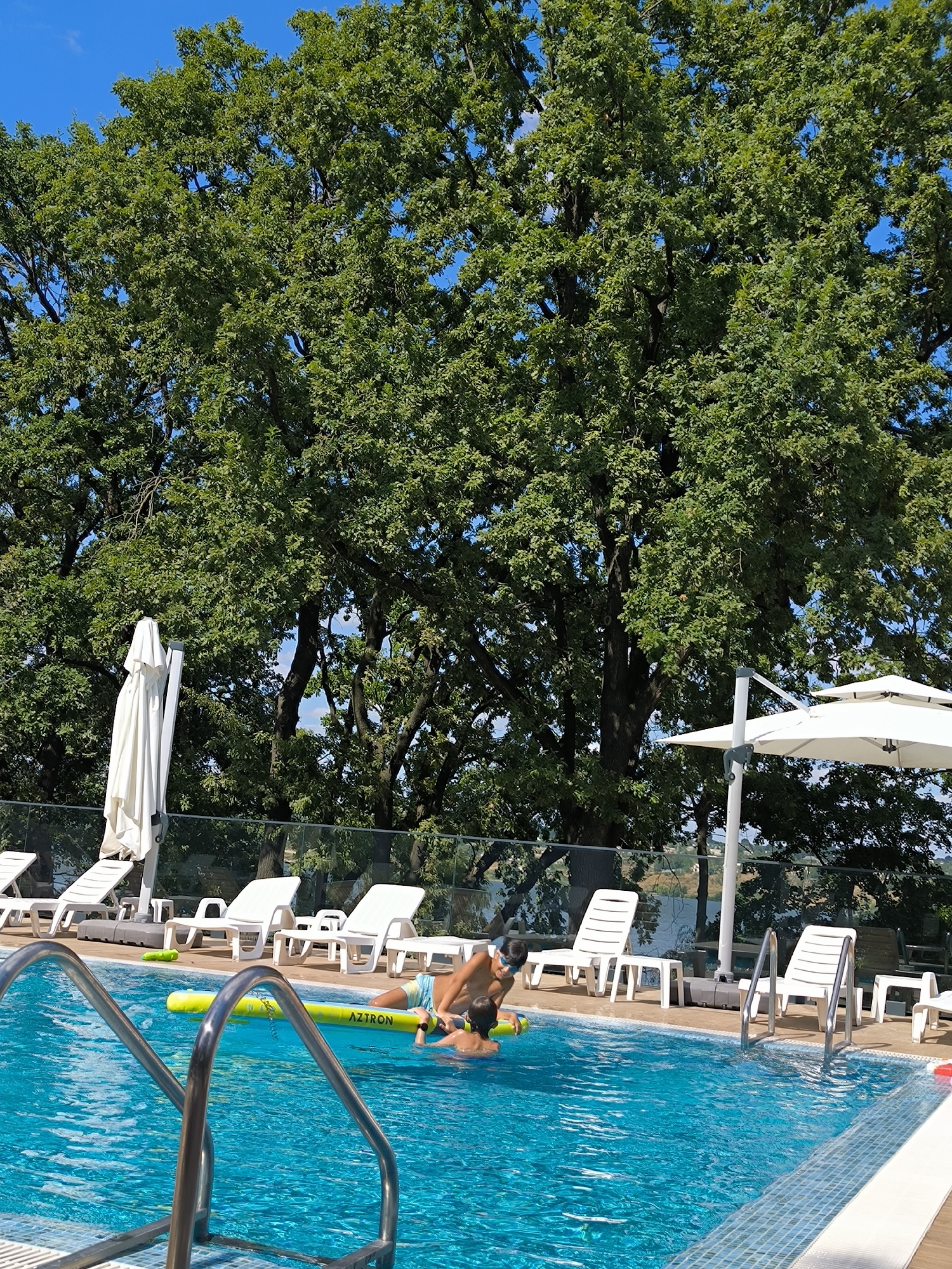 Two young boys are in a bright blue swimming pool, playing with a yellow inflatable pool toy. One boy, wearing swim trunks, is on the toy while the other, wearing goggles and swim trunks, is partially submerged in the water. The pool is surrounded by white lounge chairs and umbrellas. In the background, a dense canopy of green trees meets a clear blue sky. The scene appears to be in a resort or park setting in Ustia, Moldova, given the naturalistic backdrop of lush vegetation and clear sky, typical of summer in the region. The overall impression is one of a recreational outdoor activity on a sunny day.