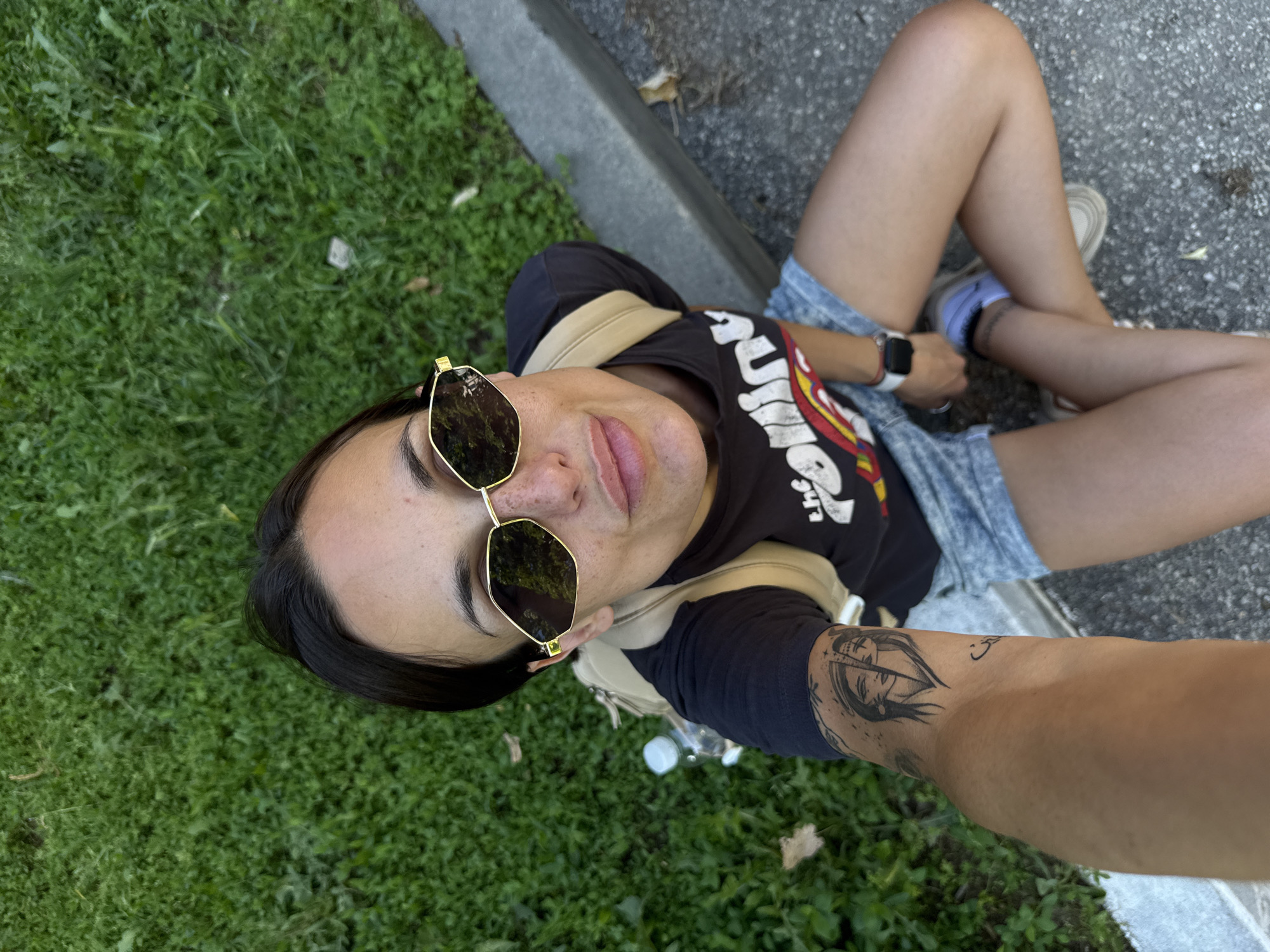 A female subject is captured in a high-angle outdoor selfie, seated on the ground in a public area of Pleven, Bulgaria. She has dark hair, is wearing gold-framed hexagonal sunglasses, and exhibits freckles on her visible facial skin. Her lips are slightly pursed. Her attire includes a black t-shirt with a partial white and red graphic print, light blue denim shorts, white socks, and light-colored athletic shoes. A beige backpack is positioned on her back. She wears a white Apple Watch with a black band on her left wrist. A black ink tattoo depicting a split face, with one half appearing female, is visible on her right forearm. The immediate surroundings consist of green grass on the left, a concrete curb extending horizontally across the mid-right, and dark grey asphalt or gravel ground in the foreground and lower right. A clear plastic water bottle with a white cap lies on the grass behind the subject. Scattered dry leaves are present on both the grassy and paved surfaces.