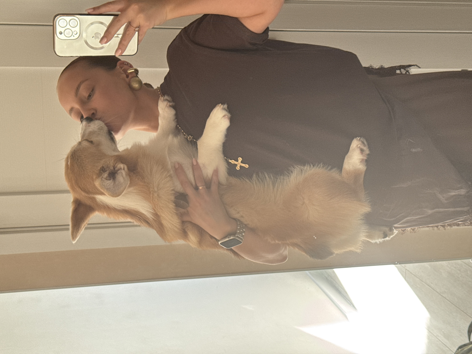 A woman is holding a corgi dog and kissing it. She is wearing a brown dress and a gold cross necklace, as well as a watch. She is also holding a phone in her left hand to take the picture. The location appears to be indoors, possibly a home. The image was not taken in the city of Chaikovsky, Russia.