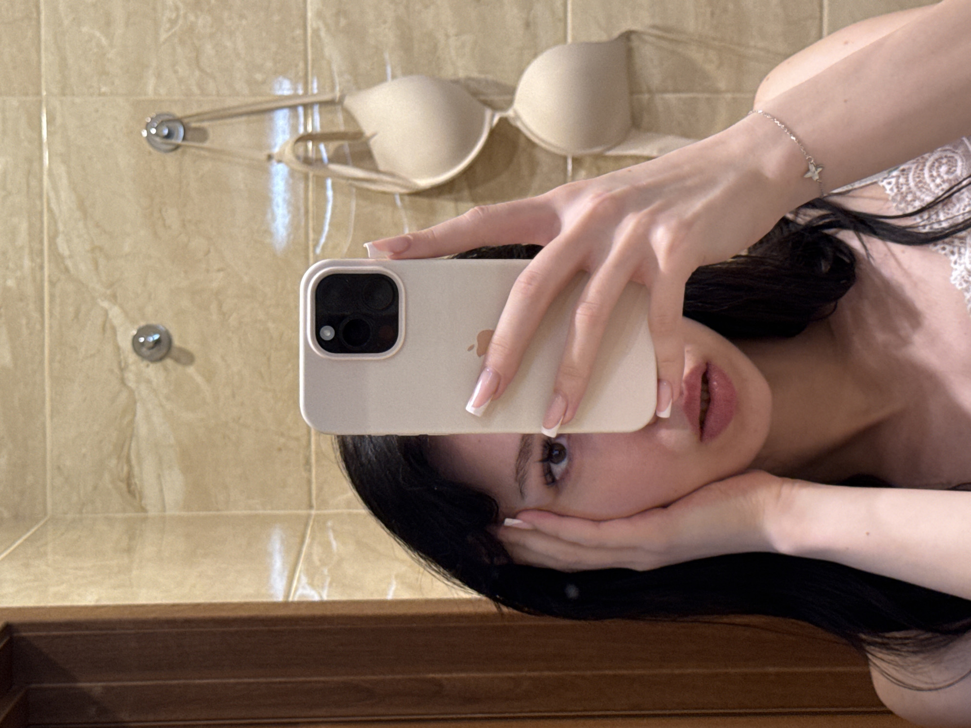 The image shows a young woman with dark hair taking a mirror selfie in a bathroom. She is holding a smartphone in her right hand, which partially obscures her face. She is wearing a camisole with lace detailing. In the background, a beige bra is hanging on a hook on a tiled wall. The overall environment suggests a residential bathroom. There is no specific indication of the location being in the city of Khimki, Russia.