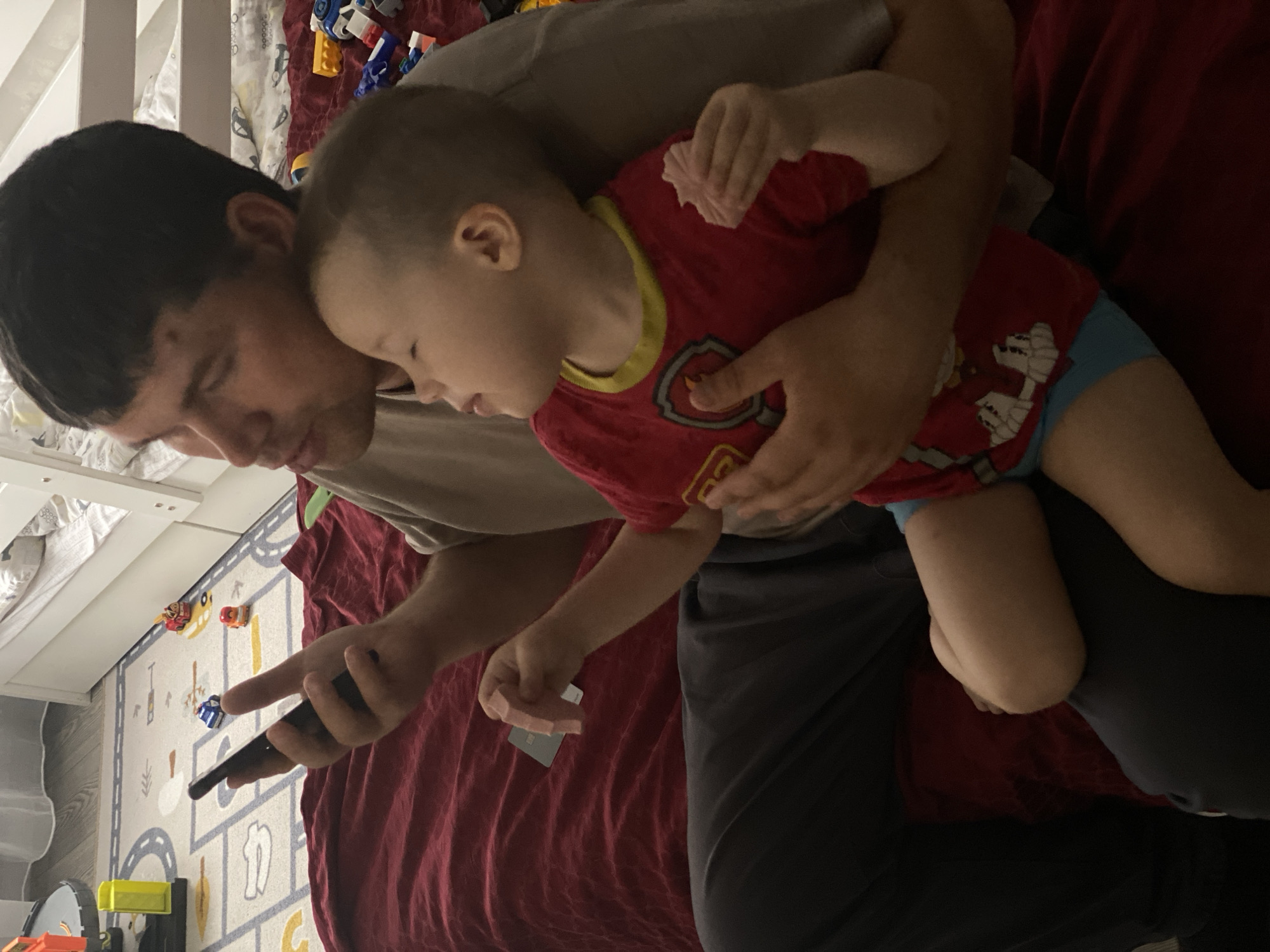This image, captured in the city of Sochi, Russia, depicts two individuals in an indoor domestic setting. An adult male, with short dark hair and wearing a light-colored t-shirt and dark trousers, is seated on a dark red blanket. He holds a dark-colored smartphone in his right hand. His left arm is positioned around a young child. The child, with light brown hair and wearing a red t-shirt with a yellow collar (featuring a visible Paw Patrol badge) and light blue shorts, is leaning against the adult male's chest. The child's eyes are closed, and its mouth is slightly open. In its left hand, the child holds a light pink, fluted, possibly edible object, with a smaller piece also visible in its right hand. A dark-colored card rests on the red blanket next to the child's left arm. The background reveals a light-colored floor rug patterned with roads and arrows, upon which various toys are scattered, including a grey and orange multi-level toy garage and several small toy vehicles (blue, red, orange). A white wooden bed frame with white bedding is visible on the right, with additional scattered building blocks nearby. Partially visible sheer white curtains are present in the upper left background.
