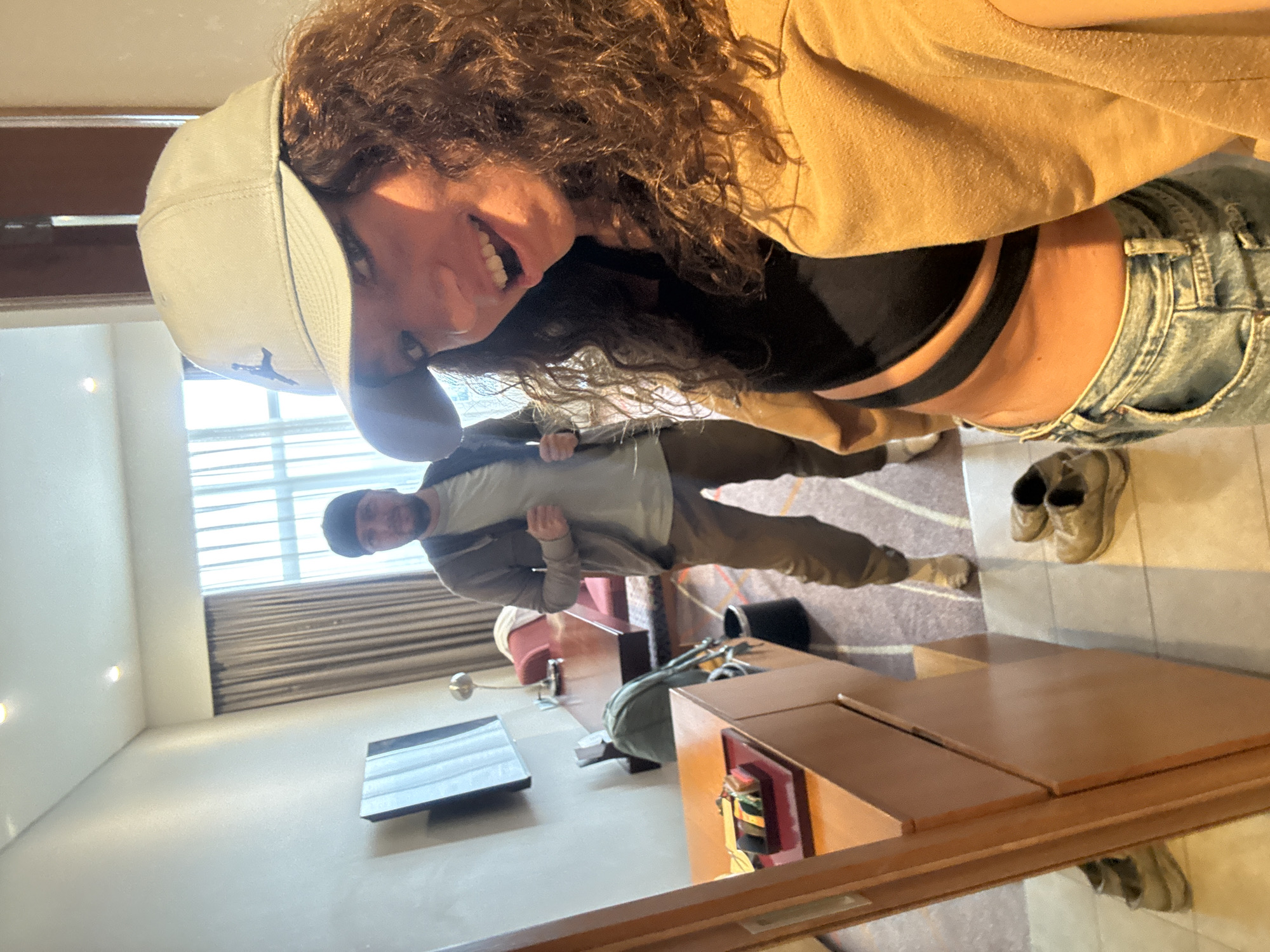The image shows two people in a hotel room. A woman with curly hair and a baseball cap is holding the camera, taking a selfie. She is wearing a tan jacket, a black top, and blue jeans. Behind her stands a man in a gray shirt, brown pants, and a brown jacket. The room features a bed, a TV, luggage, a desk, and a window with blinds. Shoes are visible on the floor near the desk. Based on visual similarities and the possible location inferred from context (e.g., tourist attire and hotel setting), the room may be located in Đà Nẵng, Vietnam.
