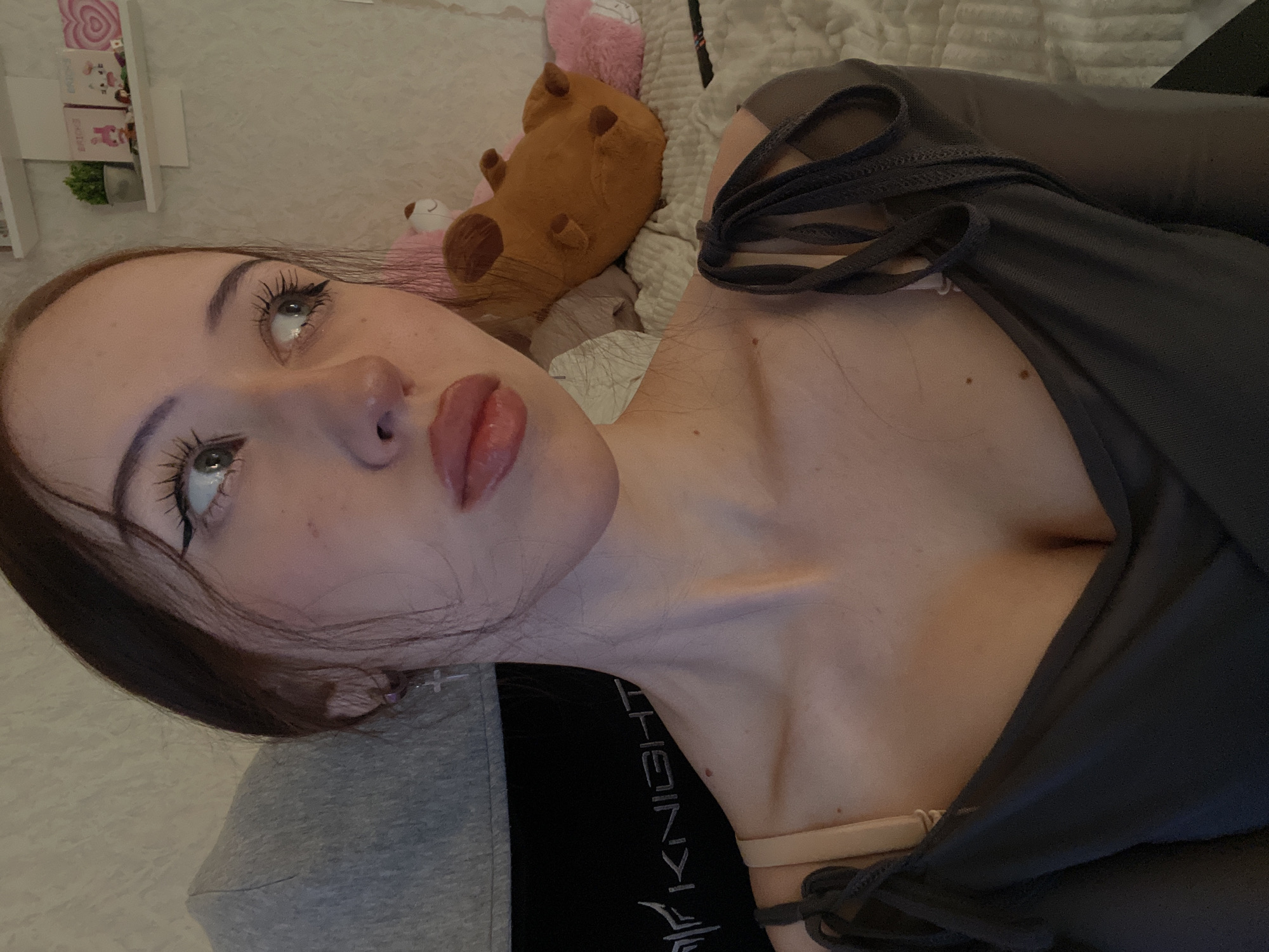 A medium-shot, high-angle portrait captures a young woman from the chest up, positioned on her right side, her head turned to look up towards the left of the frame. Her dark brown hair cascades around her shoulders. She wears a dark gray top with tie details over a light-colored bra. Her fair skin shows a few freckles and moles. Her makeup includes defined eyebrows, long dark eyelashes, and a subtle shimmer on her lips. Her eyes are a light green or hazel color.
In the background, behind her head, are various objects, including a white shelf unit with small items and a green plant, as well as several plush toys, one of which is a brown capybara and another a pink animal. A white textured fabric, possibly a blanket or comforter, is also visible. The woman's expression is neutral and contemplative. The lighting suggests an indoor setting, possibly a bedroom, with soft, even light. Part of a dark garment with the word "KNIGHT" written vertically in white is visible on the lower left of the frame. The image appears to be taken during the daytime.