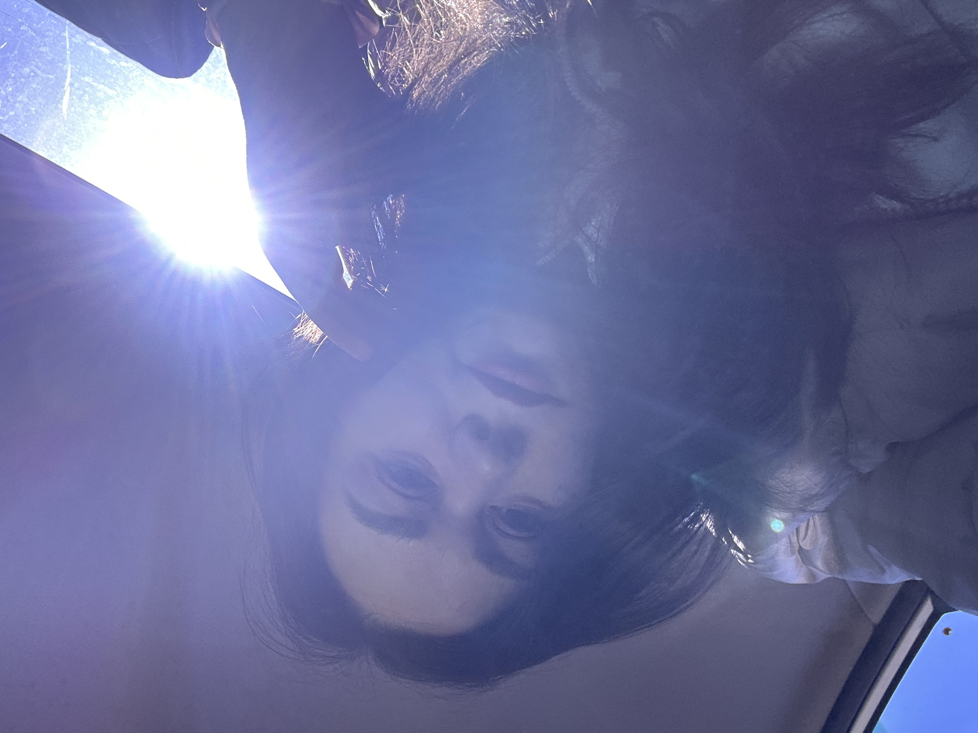 The image depicts a person with long, dark hair looking down at the camera. The lighting is very bright, with the sun shining from behind the person. The background appears to be a car interior, suggesting the photo was taken inside a vehicle. There is no indication of the location being Chaikovsky, Russia.
