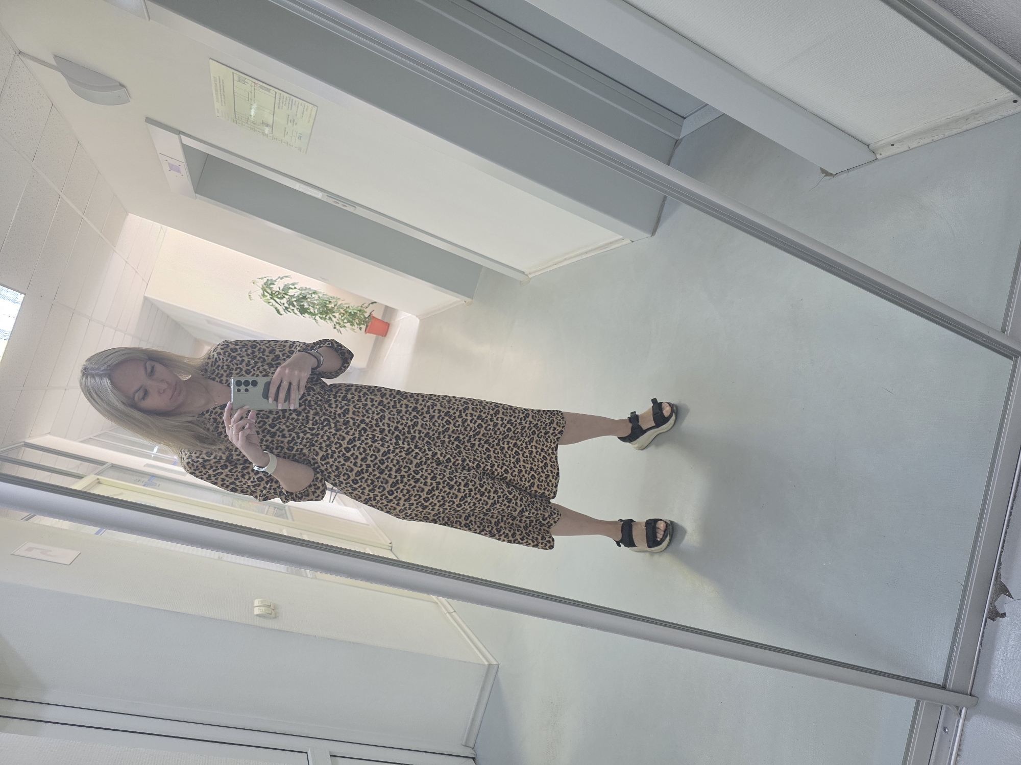A woman is taking a mirror selfie in a hallway. She is wearing a leopard print dress and black sandals. The hallway is white and well-lit, with a plant visible in the distance. The city is Chaikovsky, Russia.
