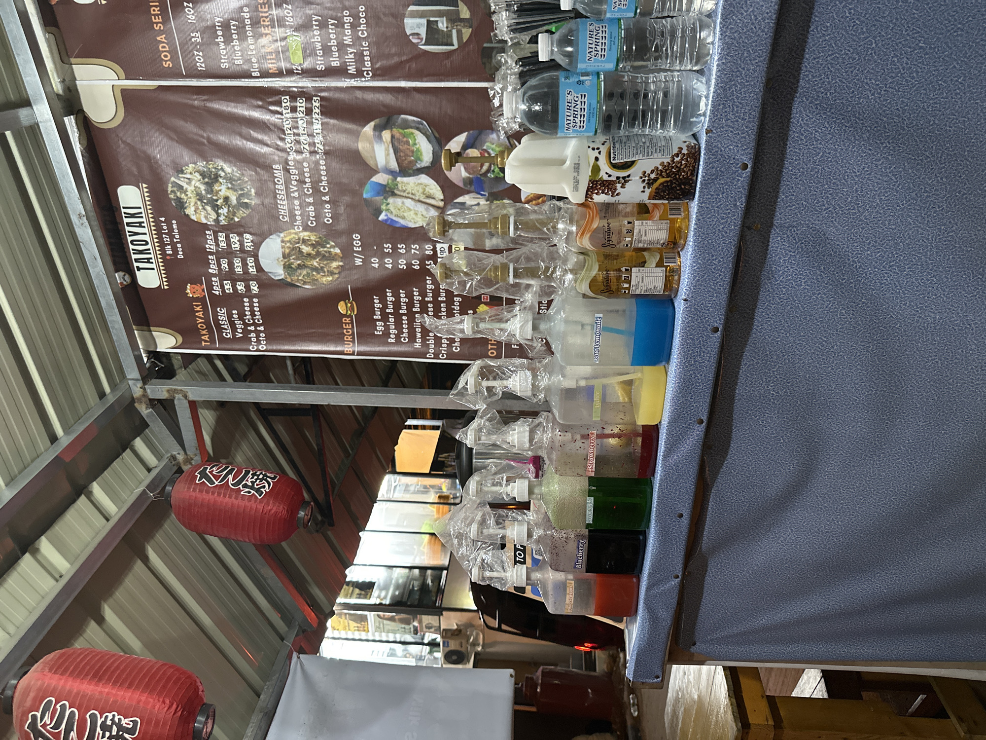 This media file captures a well-stocked food and drink stall, likely in an outdoor or semi-outdoor setting, possibly a night market or food park in Davao City, Philippines, given the address "Blk 127 Lot 4 Deca Talomo" on the banner. The stall is set up for commercial food service, with no people visible in the frame.
The central subject is a blue-grey textured counter laden with various beverages and ingredients. On the counter, numerous bottles of "Nature's Spring" purified water are stacked, alongside an array of clear plastic pump bottles containing colorful syrups, labeled with flavors such as "Strawberry," "Lychee," "Green Apple," "Blueberry," and "Blue Lemonade." Larger "Signature" brand syrup bottles and a white dispenser are also present. The tops of many syrup bottles are covered with plastic bags, suggesting a practice of hygiene or protection.
A large brown banner, serving as a menu, hangs vertically behind the counter, listing food items and prices. Prominently displayed is "TAKOYAKI," with options like Classic, Veggies, Crab & Cheese, and Octo & Cheese, available in 4-piece, 8-piece, and 12-piece servings. Other menu sections include "CHEESEBOMB" and "BURGER" (featuring Egg, Regular, Cheese, Hawaiian, Double Cheese, Crispy Chicken, and Cheesy Hotdog options, with or without egg). The banner also lists "SODA SERIES" and "MILK SERIES" with flavors like Strawberry, Blueberry, Blue Lemonade, Milky Mango, and Classic Choco, in 12oz and 16oz sizes.
Two red lanterns adorned with white Japanese characters hang from the metal roof structure overhead, contributing to an Asian-themed ambiance. The lighting suggests it might be evening or nighttime, enhancing the food stall atmosphere.