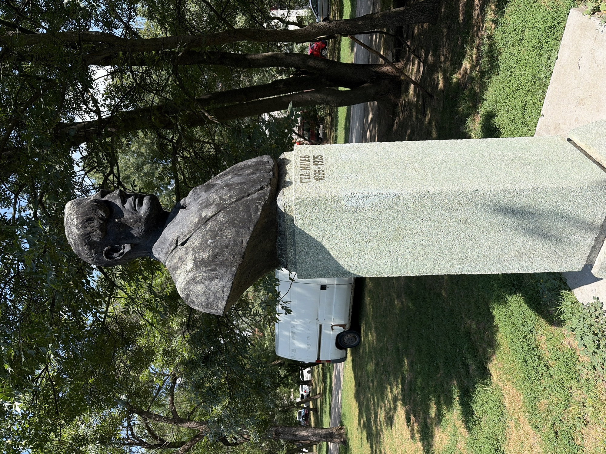 This is a bust of Geo Milev located in Sofia, Bulgaria. The bust is made of dark metal and depicts a man from the chest up, facing to the left with his head slightly down. He is wearing a suit jacket and shirt. The bust is mounted on a light green, rough-textured stone pedestal. Inscriptions are visible on the front of the pedestal, appearing to be in Cyrillic letters and numbers: "ГEO. МИЛЕВ" and "1895-1925".
The bust is situated outdoors on a sunny day, with dappled sunlight filtering through the leaves of mature trees that surround it. The ground is covered in green grass, with some patches of dry, brown grass. In the background, a white panel van is parked on a paved area, and a few other vehicles are visible further in the distance. The environment appears to be a park or a quiet street.