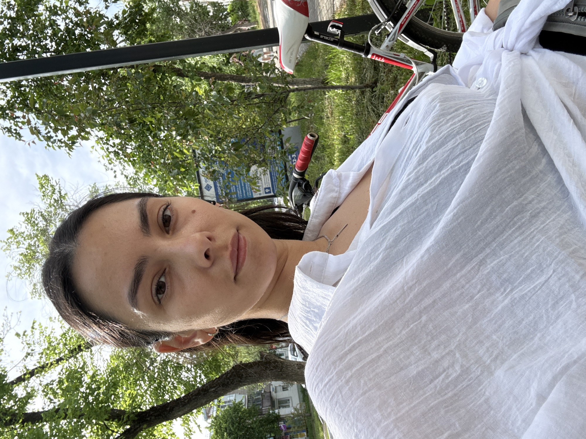 A woman is taking a selfie outdoors. She has long dark hair, fair skin, and is wearing a white blouse. She is positioned near a bicycle with red and white accents. The background includes trees, a building, and a sign with blue and white text. The location is Chaikovsky, Russia.