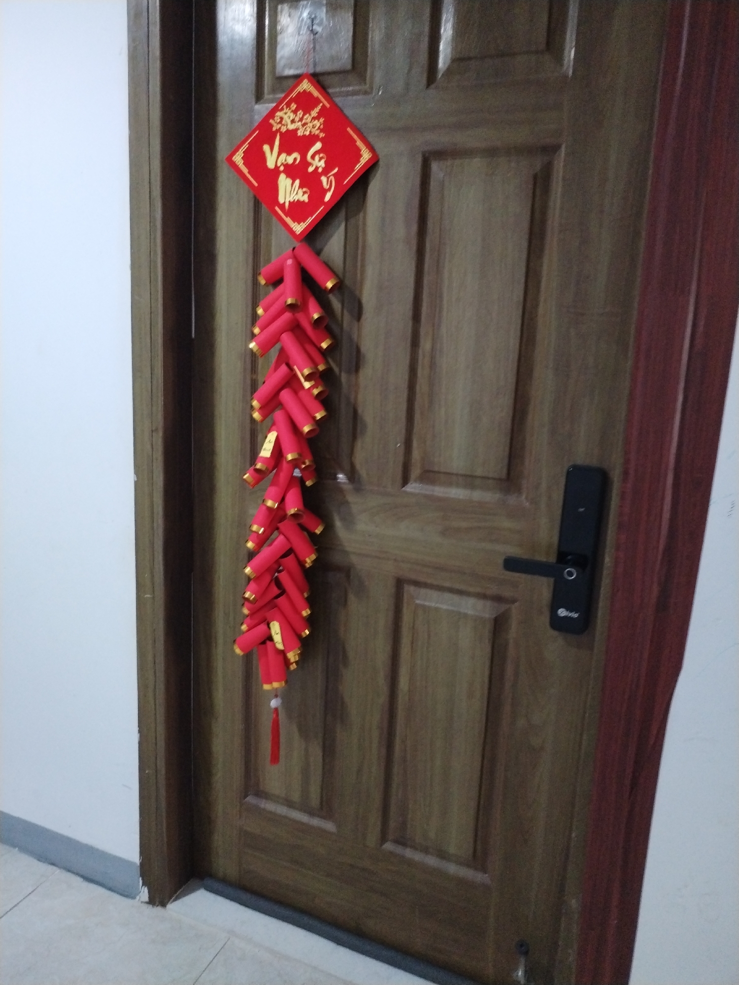 A wooden door with a dark red trim is adorned with a red diamond-shaped decoration featuring gold lettering. Below this, a string of red firecrackers with gold accents hangs down the center of the door. To the right of the firecrackers, a black electronic door lock is installed. The door is positioned in what appears to be a hallway, with a white wall to the left and a light-colored tiled floor at the bottom of the frame. The scene suggests a festive or celebratory atmosphere, common during holidays like Tet in Vietnam, and is likely located within a residential or commercial property in Nha Trang Ward, Vietnam.