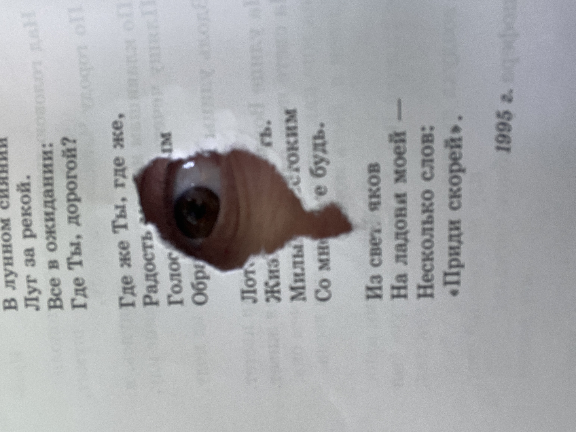 The image shows a page from a book with a hole in it, through which a human eye is visible. The text on the page is in Russian and appears to be a poem, with the date "1995 г." at the bottom. The book page is white. There are no people in the frame. It's impossible to know if the image was taken in the city of Chaikovsky, Russia, because there are no clear location markers.