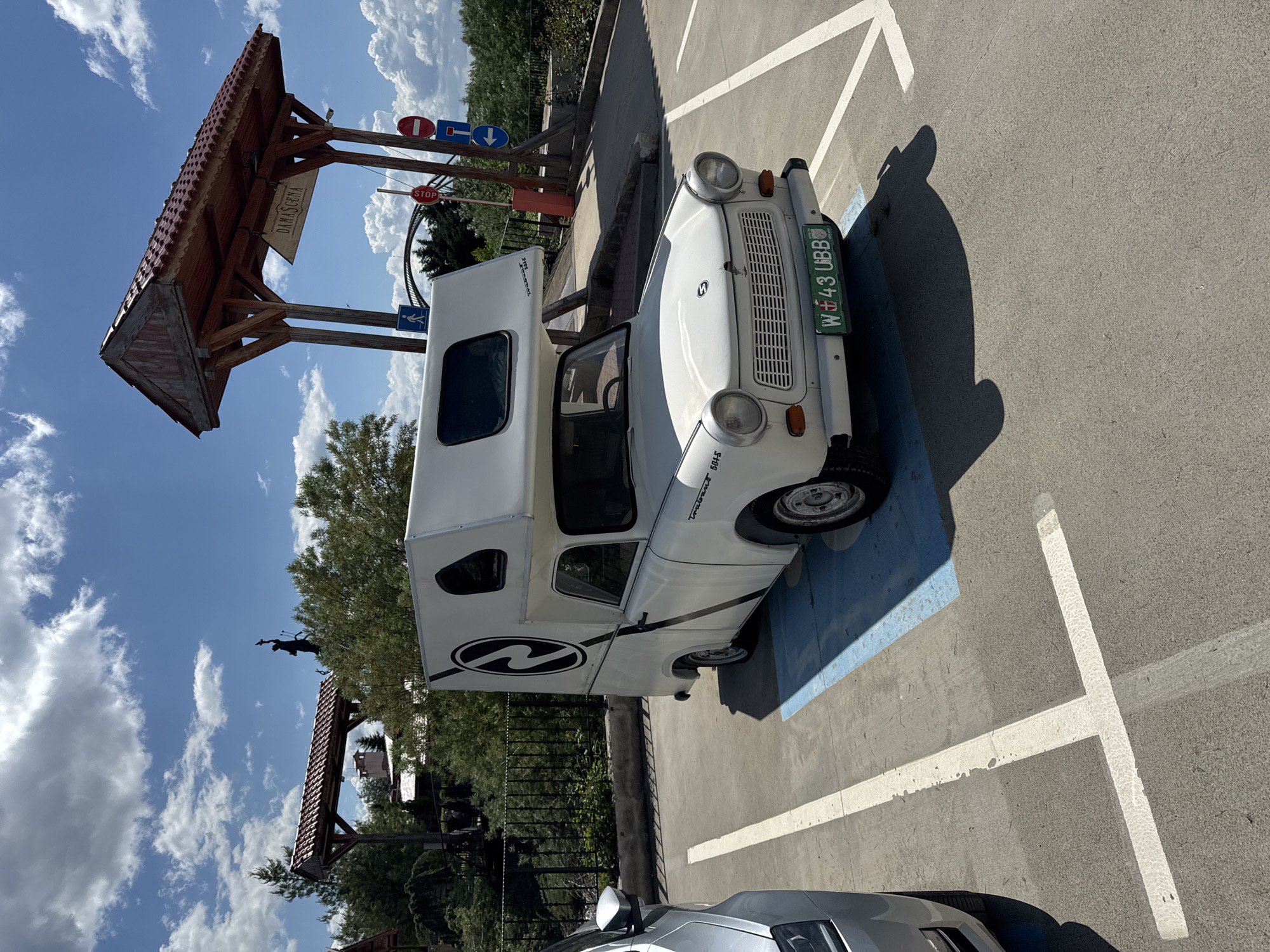 The image depicts a white Trabant 500 camper van parked in a parking space marked with blue paint, possibly indicating accessibility. The license plate, visible on the front of the vehicle, reads "W-43 UBB," suggesting registration in Bulgaria. The van is parked adjacent to a wooden archway structure with signs. In the background, partly obscured by trees, is a statue of a person on a horse. The parking lot is gray asphalt with white painted directional arrows. The sky is blue with scattered clouds. Based on the sign "DAMASCENA" visible in the background, this scene is most likely located in or around Skobelevo, Bulgaria. Another silver car is visible in the lower part of the image.