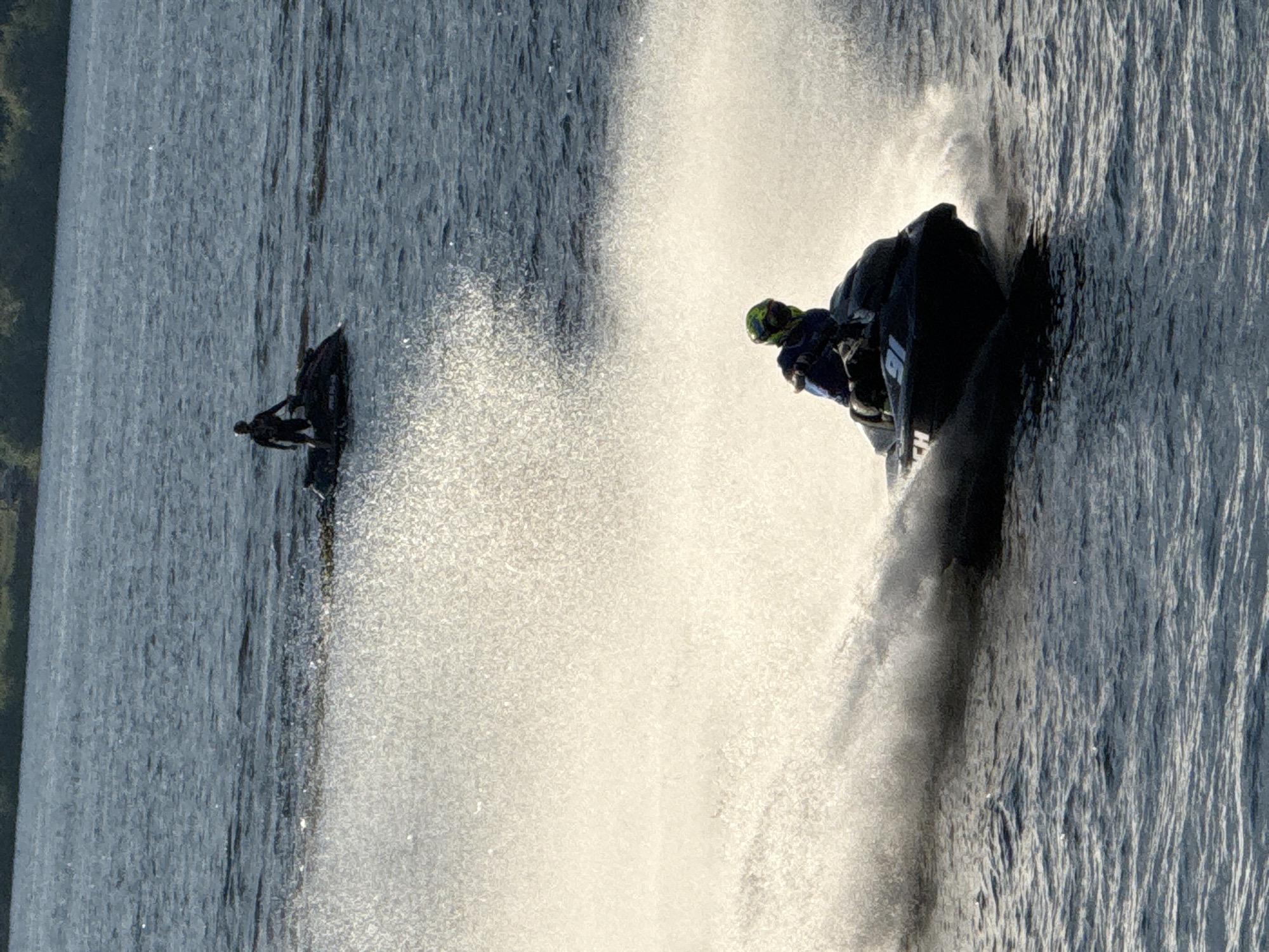 This image shows two jet skis on a body of water. The closest jet ski has a rider wearing a helmet and a blue jacket, creating a large spray of water as it moves. The jet ski has the number "91" visible on its side. Further in the background, another jet ski is visible with a rider standing on it. The water is dark blue and appears to be choppy. The background shows a distant shoreline with trees and a light-colored sky. The overall scene suggests a recreational activity on the water. The image does not contain any information to suggest it is located in the city of Perm, Russia.