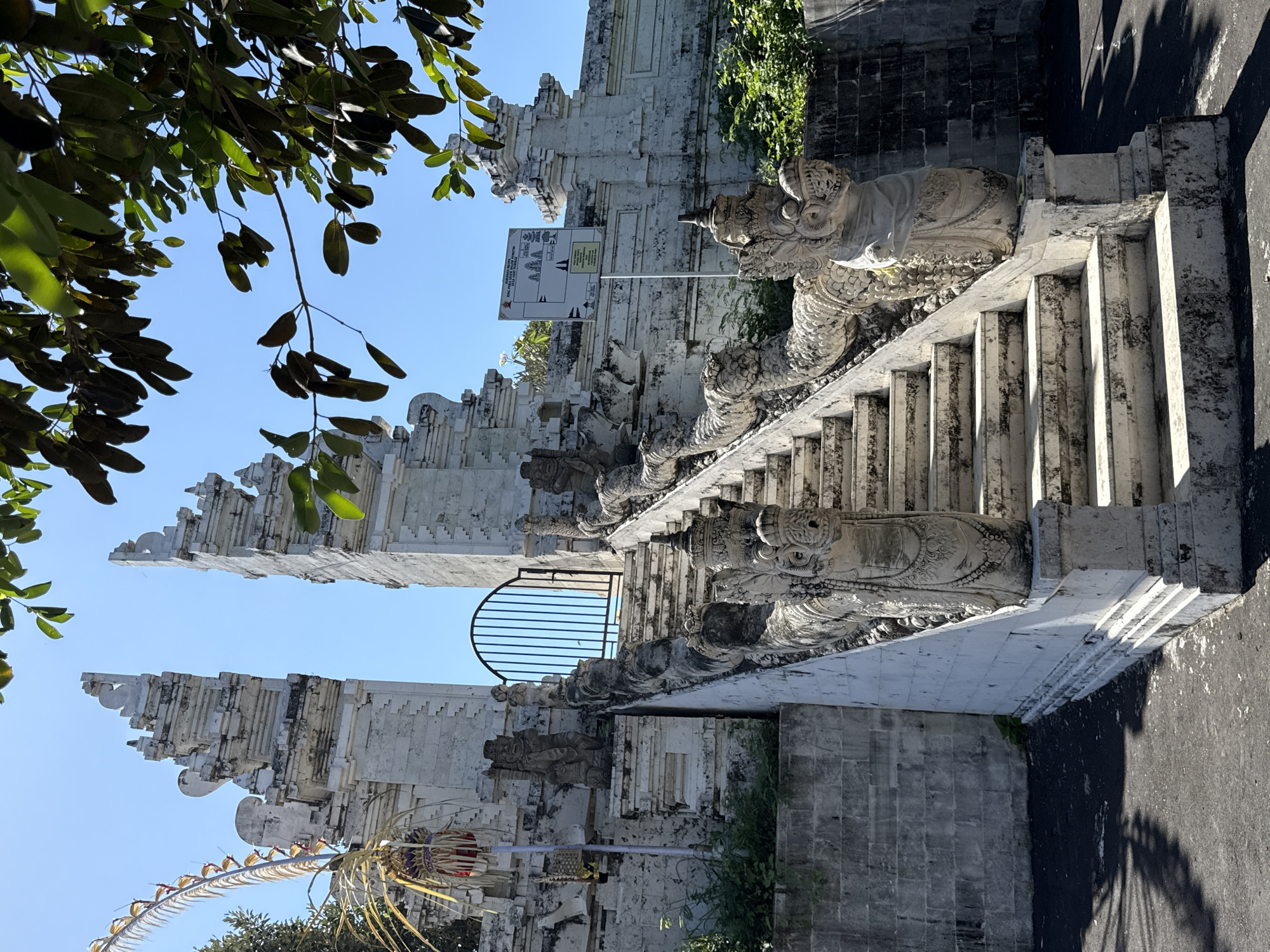 The image depicts the Pura Dalem Jenggala, a temple located in the Kuta Selatan area of Bali, Indonesia. The temple features a grand white stone staircase leading up to its entrance, flanked by ornate dragon sculptures. The staircase is a prominent feature, with intricate carvings and weathered stone that suggest age and significance. At the top of the stairs, a black metal gate stands, guarding the path further into the temple complex.
The architecture of the temple is characteristic of Balinese style, with intricately carved stone pillars and structures. Two large, imposing gate towers, also made of white stone, frame the view of the sky and the gate beyond. To the left of the stairs, a traditional Balinese "janur" decoration, a palm leaf offering, hangs, adding a touch of cultural vibrancy to the scene.
The environment surrounding the temple appears to be bathed in bright sunlight, casting distinct shadows on the ground. Lush green foliage is visible, particularly on the right side of the image, contrasting with the white stone of the temple. A signpost is mounted on a pole to the right of the stairs, displaying information about the temple, likely in Indonesian. The sky is clear and blue, indicating a sunny day. There are no people visible in the image, focusing the attention entirely on the architectural grandeur and artistic details of the temple.