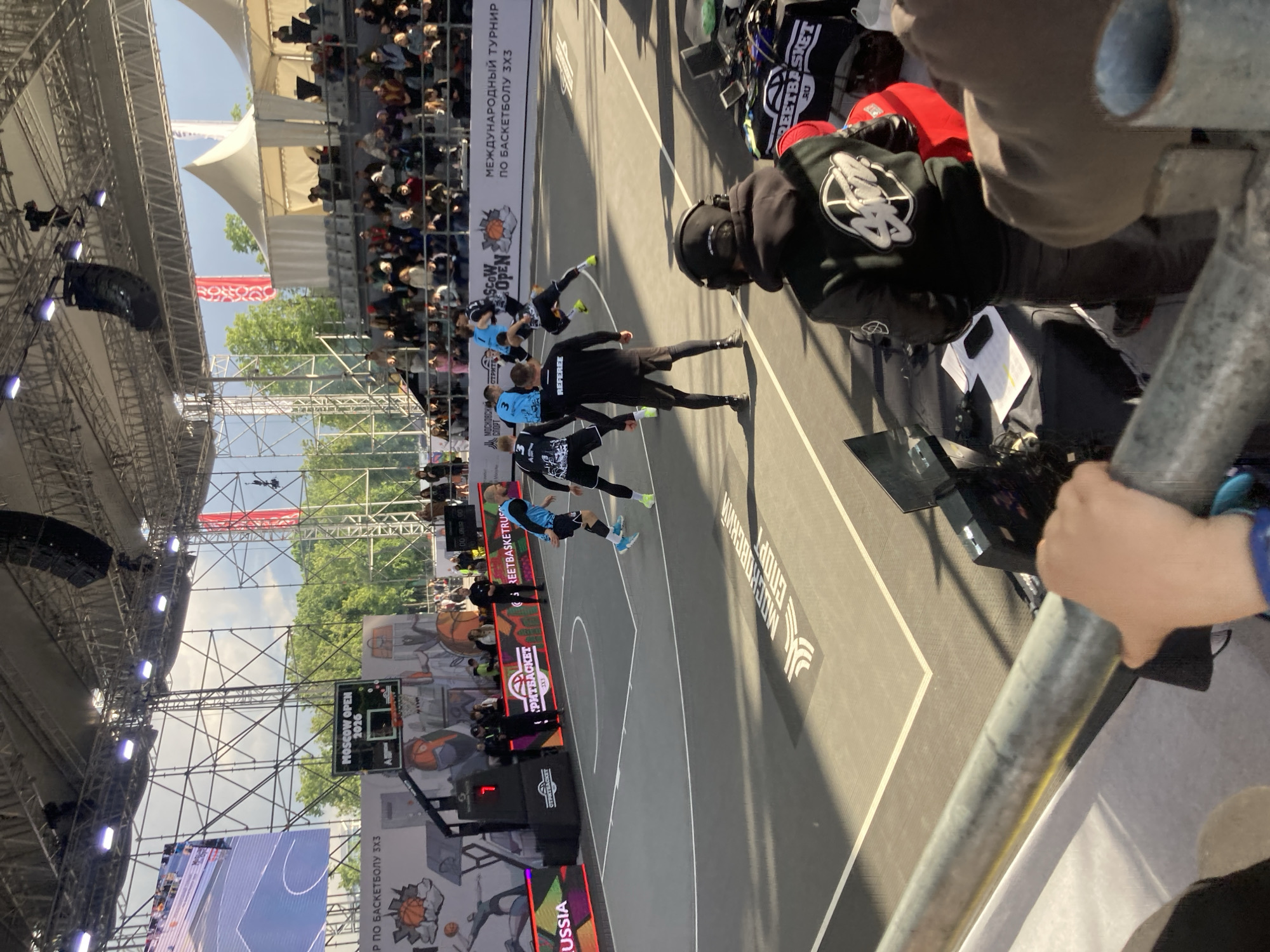 The image shows a 3x3 basketball tournament in Moscow, Russia. Players in blue and black uniforms are on a grey court. Spectators are seated in the background under a white tent-like structure. The backdrop features branding for "Moscow Open 2026", the tournament name, and other sponsorships. A referee in black stands on the court. Metal scaffolding with lights and sound equipment is overhead. A person is holding a metal railing in the foreground.