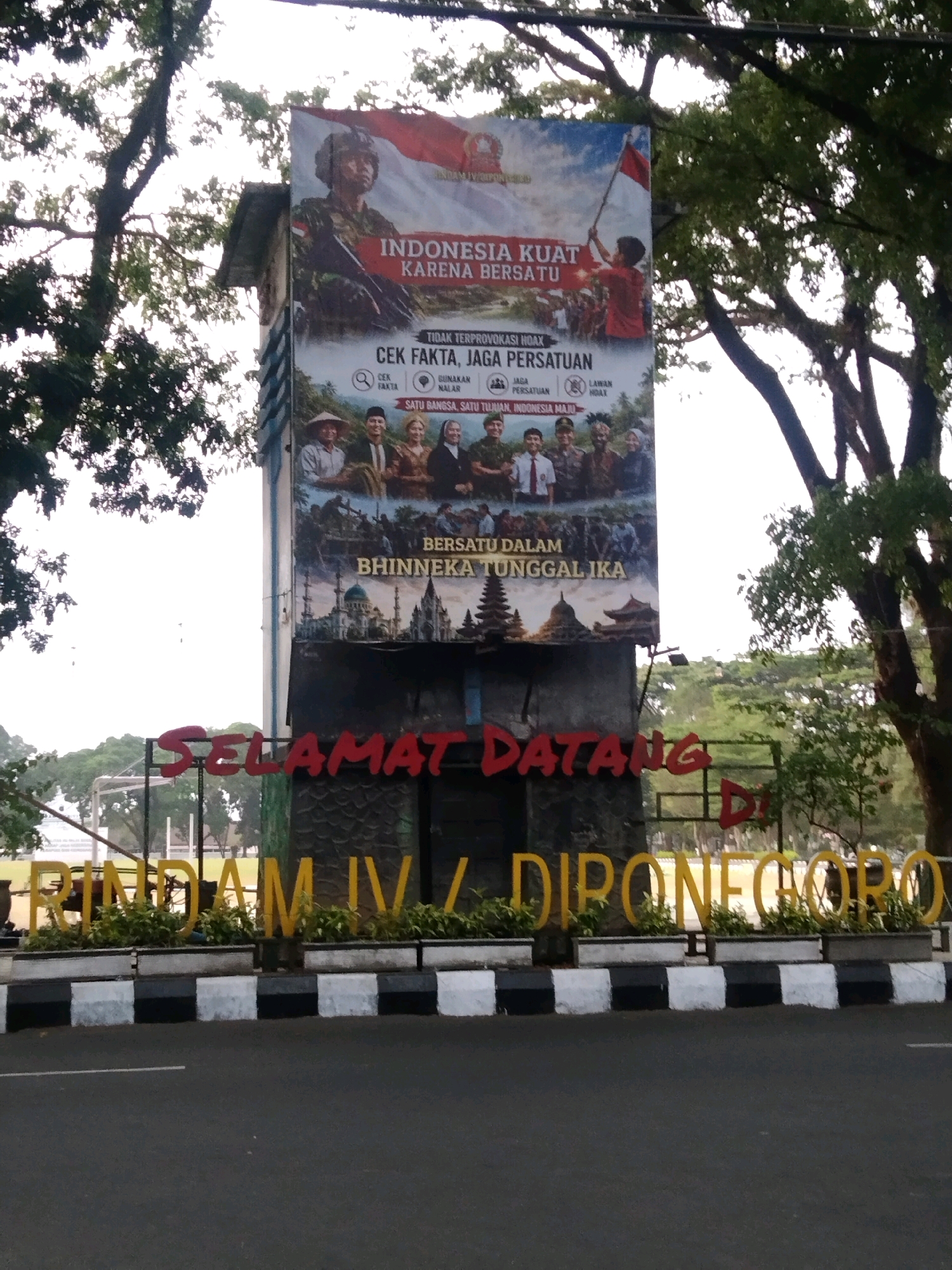 The image shows a large billboard or banner displayed on the side of a tower or monument in Magelang, Indonesia. The banner features patriotic imagery, including a soldier, the Indonesian flag, and various cultural symbols, with text promoting national unity and strength. Below the banner, large red letters spell out "SELAMAT DATANG DI", meaning "Welcome to". Further down, large yellow letters spell out "RINDAM IV / DIPONEGORO", indicating the location as Rindam IV Diponegoro, a regional military command in Magelang. The scene is outdoors, with trees and a cloudy sky visible in the background, and a road in the foreground. The monument itself is constructed from stone blocks.