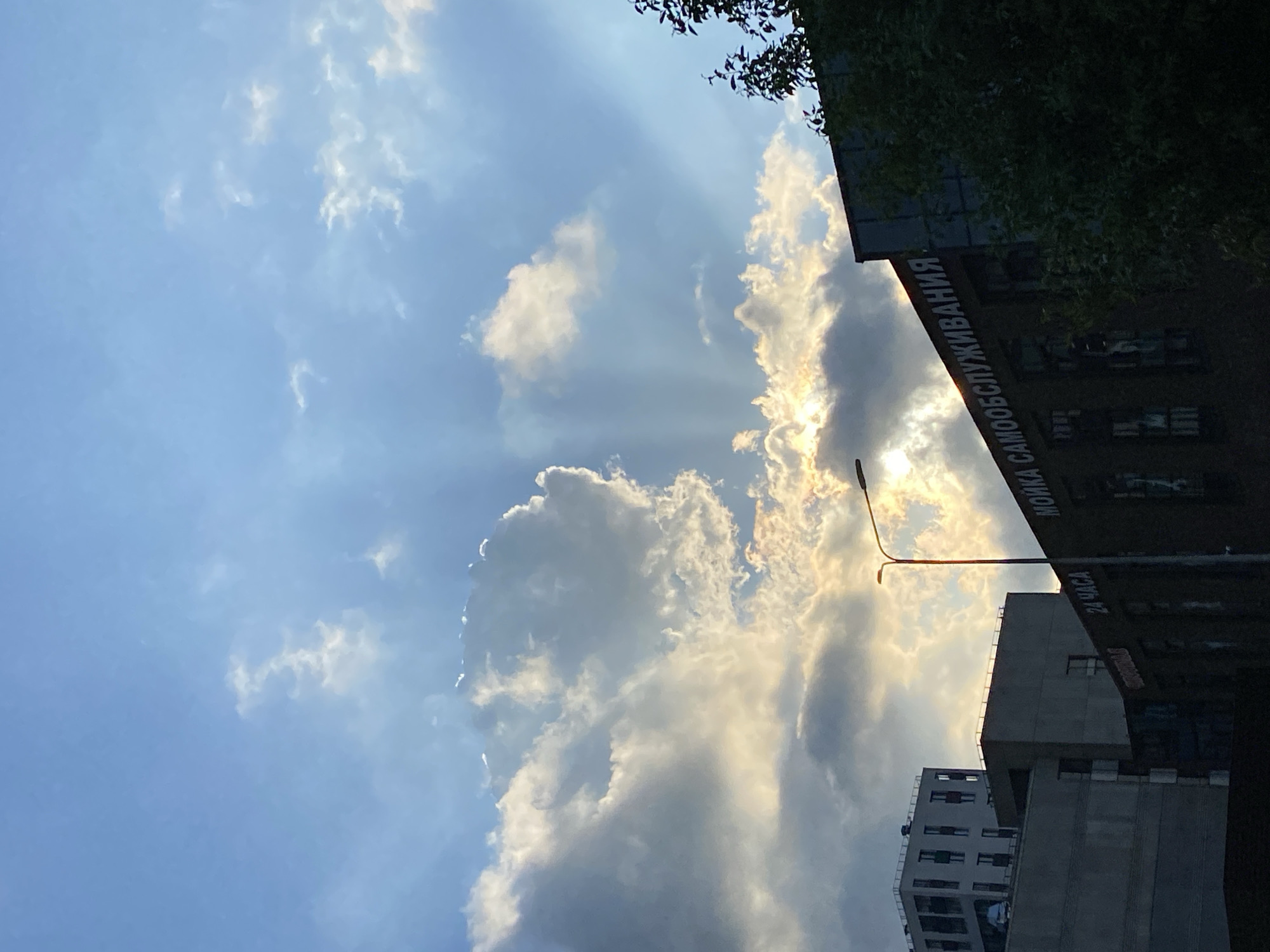In Saint Petersburg, Russia, a street lamp on a pole stands tall in the foreground, casting its light against a backdrop of buildings and a cloudy sky. The buildings are of varying architectural styles, with some featuring modern designs and others appearing older. The sky is filled with dramatic clouds, with the sun breaking through in some areas, creating rays of light that illuminate the scene. The overall impression is one of an urban landscape bathed in the soft, diffused light of a cloudy day.