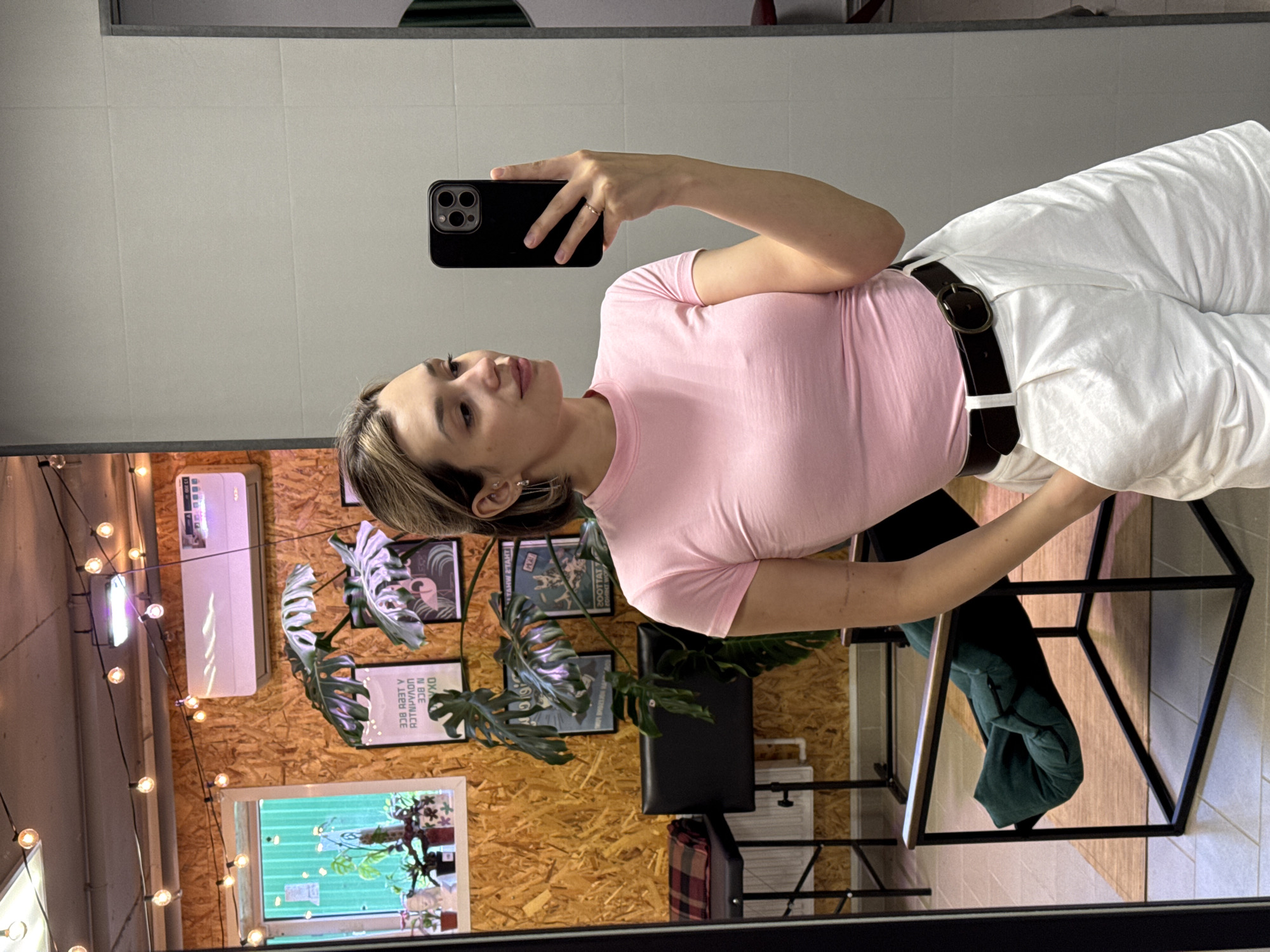 The media file is a portrait-oriented, vertically flipped selfie taken in a room with white tiled walls and a light-colored, wood-paneled accent wall. A young woman with blonde-streaked brown hair is visible, holding a smartphone in her right hand to take the picture. She is wearing a short-sleeved, light pink t-shirt and high-waisted, wide-leg white pants with a black belt. Her left arm is bent, resting on what appears to be a chair with a geometric black metal frame.
The background of the room includes a white air conditioning unit mounted on the wood-paneled wall, along with several framed pictures and plants. String lights with visible bulbs hang across the upper portion of the wood paneling. A window or mirror reflecting plants and a green wall is also visible. The lighting appears to be artificial, suggesting an indoor setting, possibly a cafe or a lounge area. The woman's expression is neutral, and the overall mood seems casual and relaxed. No specific time of day or weather conditions can be inferred. There is no visible text in the image.