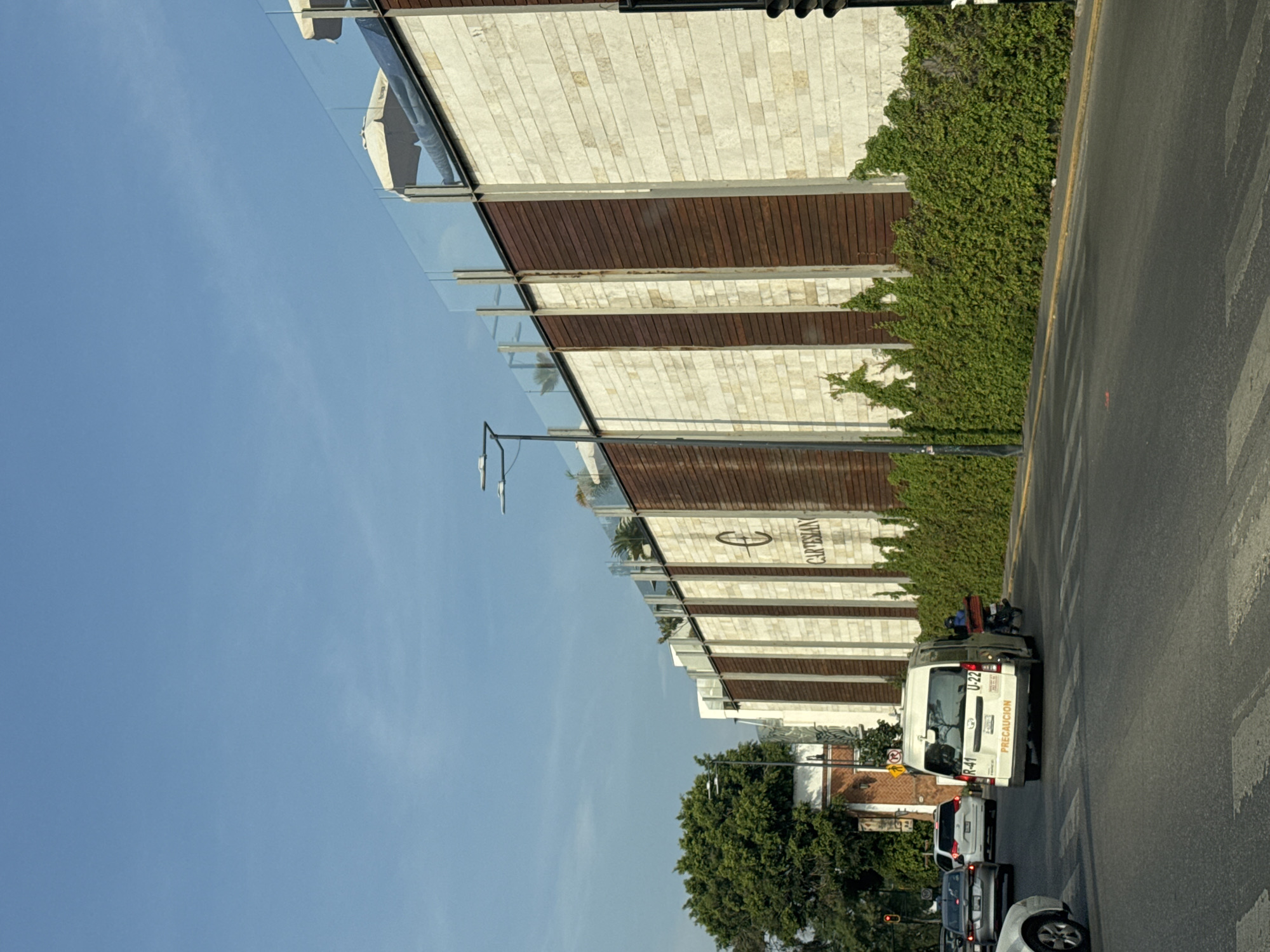 The image captures an urban street scene under a clear blue sky during daylight hours, likely in Puebla, Mexico, based on context.
To the left, a modern multi-story building features a facade constructed from alternating sections of light-colored stone bricks and dark horizontal wooden panels. Balconies or terraces with clear glass railings extend from the building. Green ivy-like vegetation grows along the base and lower sections of the building's wall. Prominently displayed on the building's facade is a logo incorporating a stylized 'C' and the word "CARTESIAN."
To the right, a paved road stretches into the distance, marked with white pedestrian crosswalk stripes and a yellow dividing line. Several vehicles are present on the road, indicating traffic. In the mid-ground, a white public transport van (likely a "combi" or "microbus") is visible from the rear, displaying the route numbers "U-22" and "R 41," along with the word "PRECAUCION" (caution). Behind this van, a green vehicle, possibly another service or taxi van, has luggage or items secured on its roof. Further back, a white SUV and a dark sedan are part of the traffic flow. The front wheel and a portion of the bumper of another white car are visible in the immediate foreground on the right.
Lush green trees are scattered throughout the background, adding natural elements to the urban landscape. A tall street light pole stands between the building and the road. The bright lighting suggests it is mid-day or afternoon.