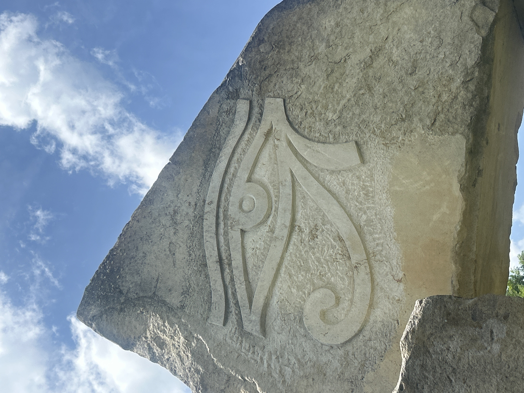The image presents a low-angle, close-up view of a large, light-colored stone monument or sculpture, situated outdoors under a clear blue sky. The primary subject is a significant portion of a roughly cut stone slab, which features an intricate, stylized design deeply carved into its surface.
The carving consists of flowing, curvilinear lines and shapes, forming an abstract or symbolic motif. It appears to be a relief carving, with contours that create distinct shadows and highlights, indicating it is bathed in strong sunlight. The stone itself is textured, varying from smooth, carved areas to rougher, natural surfaces. Its color is a light grey or beige.
The background reveals a bright, clear blue sky dotted with scattered, wispy white clouds. The lighting suggests it is daytime, with direct sunlight illuminating the stone and casting shadows. No people or discernible text are visible in the frame. The scene conveys a sense of natural artistry and permanence, likely an outdoor art installation or memorial in Gardevtsi, Bulgaria.