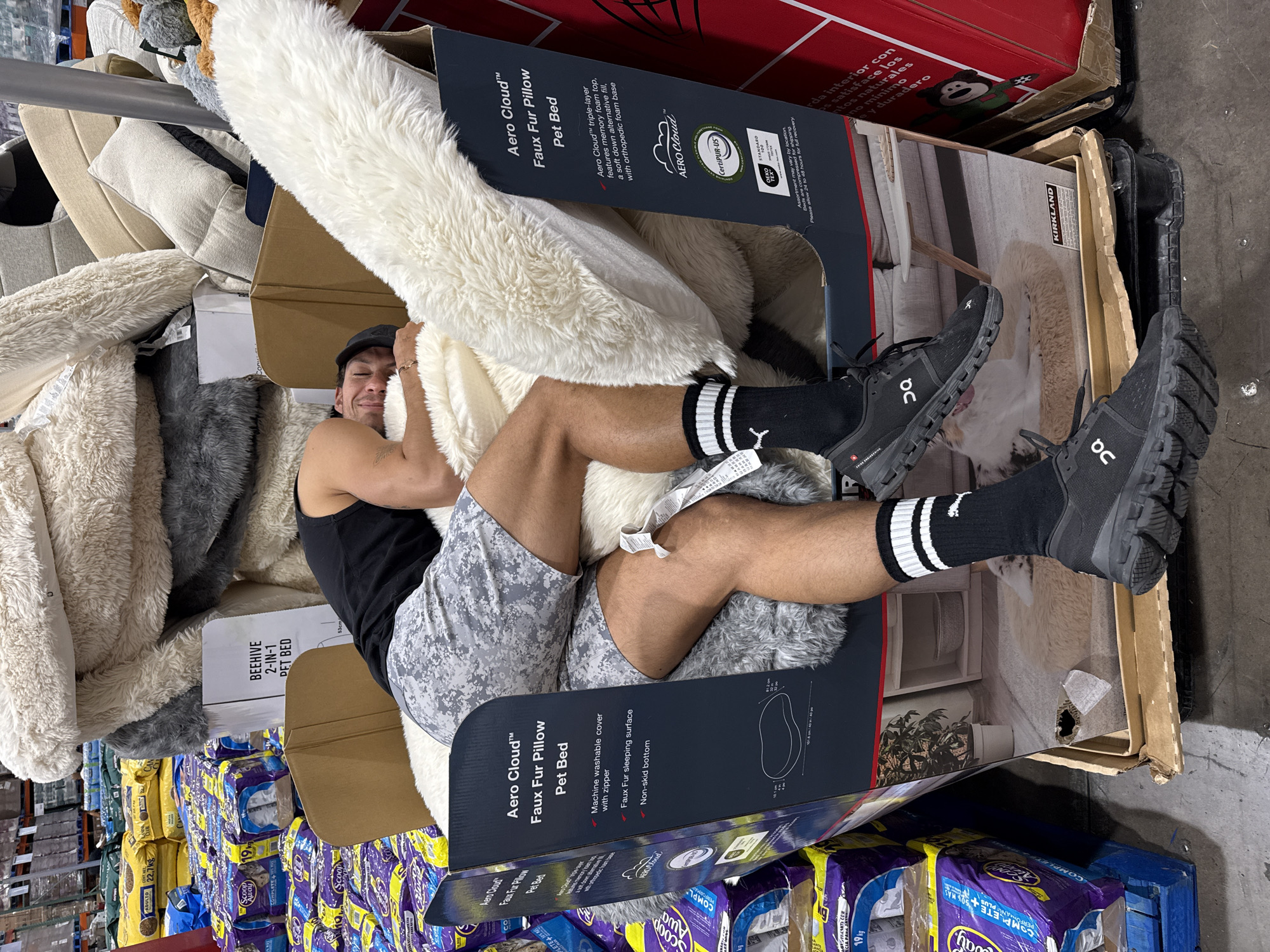 In an indoor retail warehouse setting, likely in Puebla, Mexico, a young man is playfully nestled inside a large cardboard box for an "Aero Cloud™ Faux Fur Pillow Pet Bed." He is smiling at the camera, wearing a black tank top, grey camouflage shorts, black socks with white stripes, and black athletic shoes (ON Cloud brand).
The man is surrounded by numerous pet beds, primarily soft, fluffy faux fur in shades of white, cream, and grey, some still packaged. The "Aero Cloud™ Faux Fur Pillow Pet Bed" box details include "triple-layer features memory foam top, a soft down alternative fill, with orthopedic foam base," and a "Machine washable cover with zipper," "Faux Fur sleeping surface," and "Non-skid bottom." Labels like "CertiPUR-US" and "OEKO STANDARD" are visible.
The background shows industrial shelving stocked with large quantities of products, including bags of "Scoop Away COMPLETE 19 kg" cat litter and "ADULTO 22.7 kg" pet food, as well as what appear to be packages of toilet paper or paper towels. A "KIRKLAND" logo is visible on another box, characteristic of a wholesale club store like Costco. Spanish text on a red box reads phrases like "rda interior con," "ps satisface los," "ntos naturales," and "e y duradero." Another product tag mentions a "BEEHIVE 2-IN-1 PET BED." The overall scene suggests a lighthearted moment within a busy commercial environment.