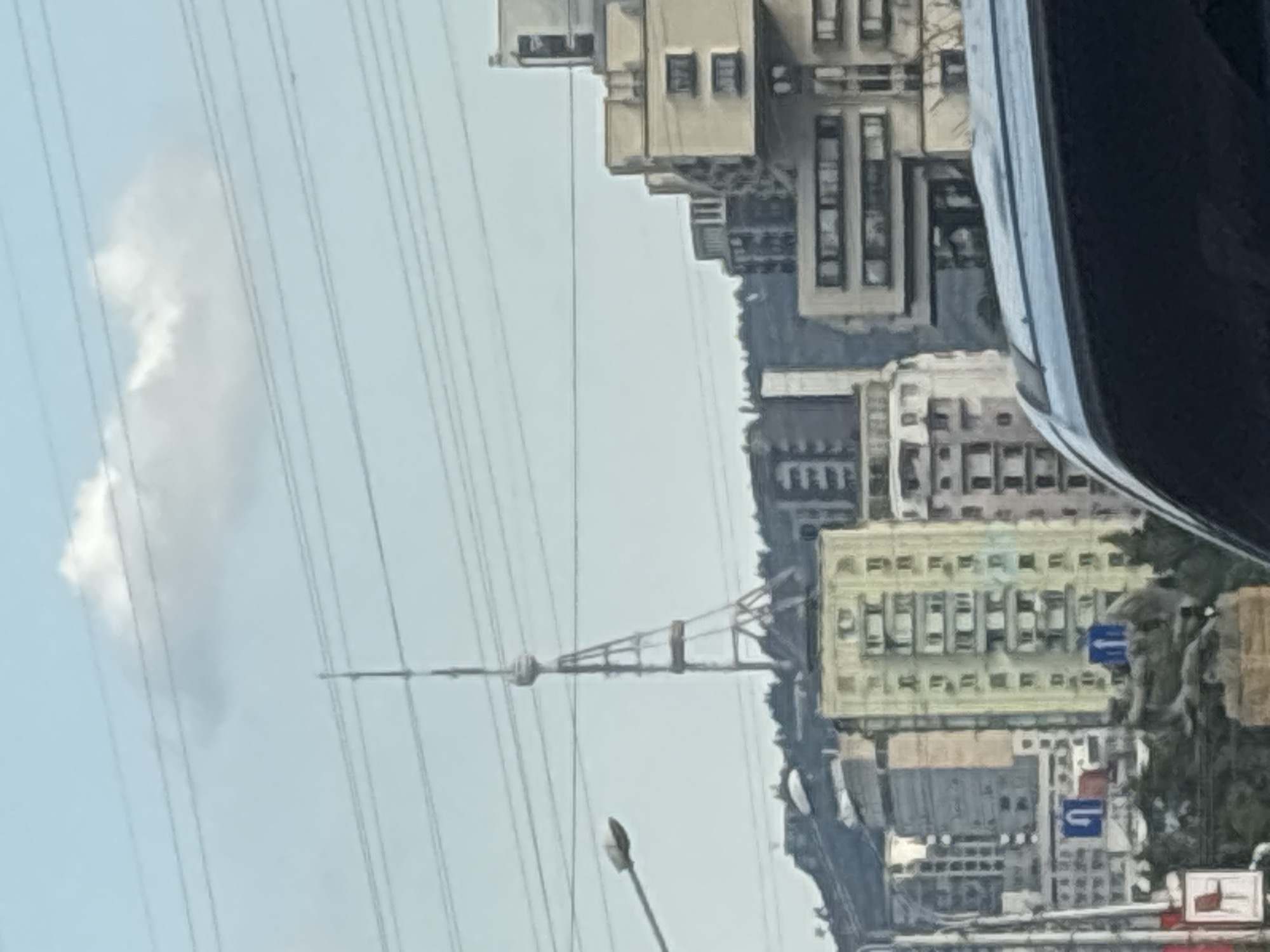 This media file captures a view of a city skyline, likely Tbilisi, Georgia. The image is oriented vertically, with the sky and power lines dominating the upper portion. The city appears to be built on a slope, with various buildings of different architectural styles and heights cascading down. In the foreground, a street light is visible, suggesting an urban environment.
The most prominent object is a tall, slender tower, possibly a telecommunication or observation tower, with a distinctive structure at its top. This tower is supported by a network of power lines that stretch across the frame, creating a visual pattern.
Several buildings are visible, some are light-colored with rows of windows, while others are darker and more industrial in appearance. One building in the mid-ground stands out with its light yellow facade and regularly spaced windows. Road signs are partially visible, including one with a blue arrow indicating direction.
The weather appears to be overcast, with a single, wispy cloud in the sky. The overall impression is of a slightly hazy, daytime scene in a densely built urban area. There is no clear indication of people or specific events taking place, but the presence of traffic signs and streetlights points to an active city.