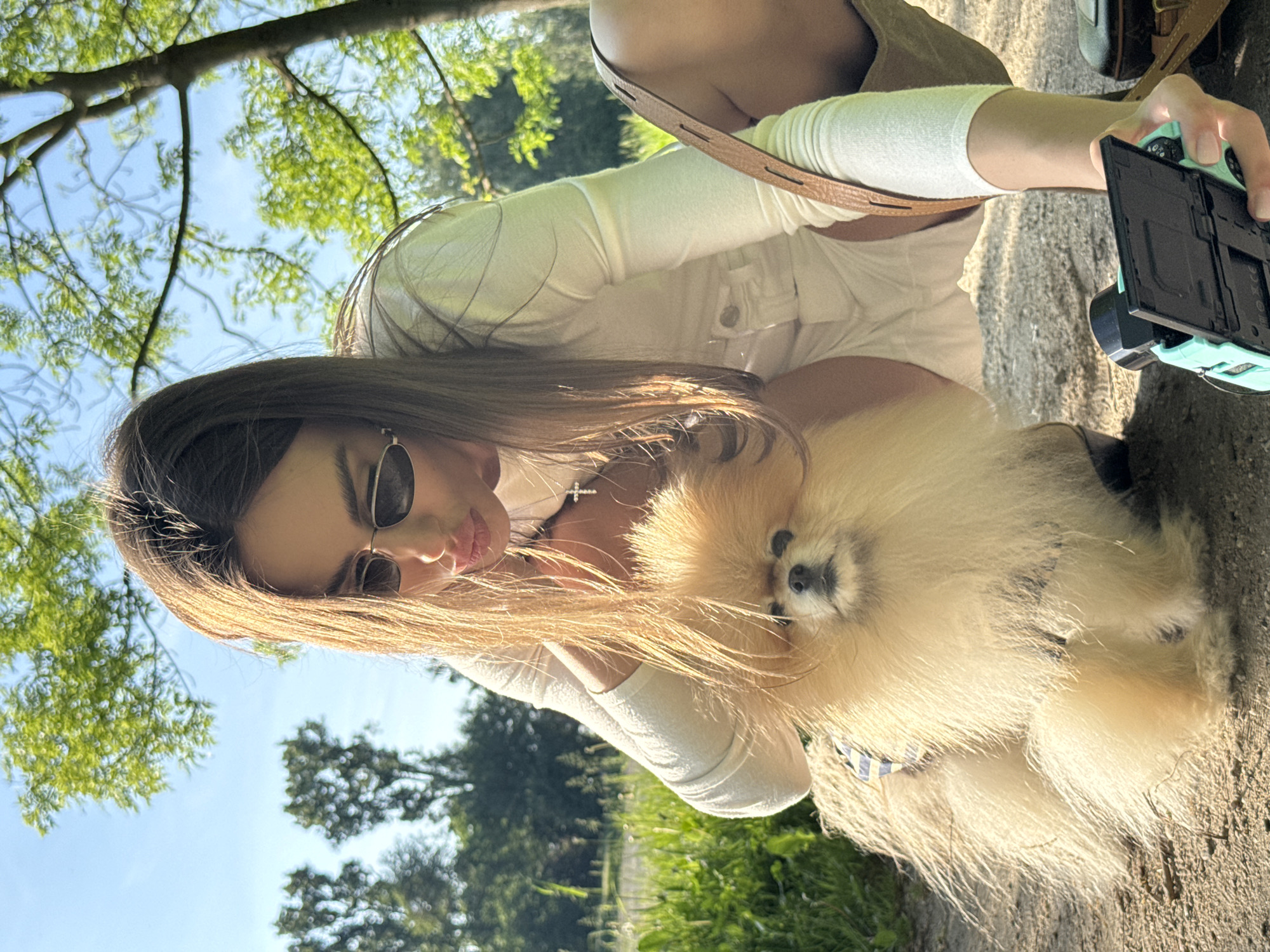 A young woman holds a small Pomeranian dog while seemingly taking a selfie or photo with a small, light blue and black camera. The woman wears a white long-sleeved shirt, white shorts or pants, sunglasses, and a delicate silver cross necklace. She kneels on a dirt path lined with grass, under the shade of trees. The dog is fluffy and light brown. The location appears to be a park or wooded area.