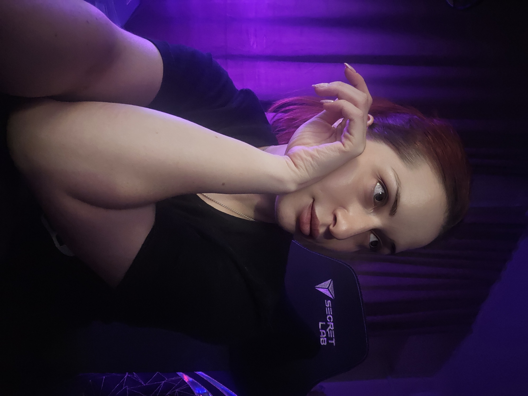 The image shows a close-up of a woman with reddish-brown hair, looking off to the right. She is wearing a black t-shirt and has her left hand resting on her cheek. Behind her, a purple curtain is visible, and she is seated in a black chair with the "SECRET LAB" logo on it. The overall lighting is dim with a strong purple hue, likely from ambient lighting. The scene appears to be indoors. No specific location in Burgas, Bulgaria, can be identified from the image.