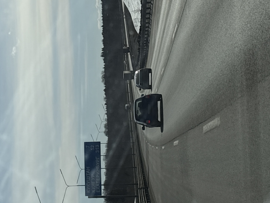 An image taken from a moving vehicle captures a multi-lane highway near Yanychi, Russia, under a partly cloudy sky. The highway, paved with asphalt, features white dashed lane markings. On the left, a large blue overhead gantry sign displays Cyrillic text: "КУКУШТАН 7", "КУНГУР 52", and "ЕКАТЕРИНБУРГ 313", indicating destinations and distances. Numerous streetlights extend along both sides of the roadway. In the immediate foreground, a dark-colored hatchback is observed in the left lane, followed by a lighter-colored sedan in the right lane, both traveling away from the camera. Further down the highway, additional vehicles, including trucks, are visible. Metal guardrails run along the median on the left and the shoulder on the right, where patches of snow are present. A dense line of evergreen trees forms the horizon, suggesting a rural or semi-rural environment.