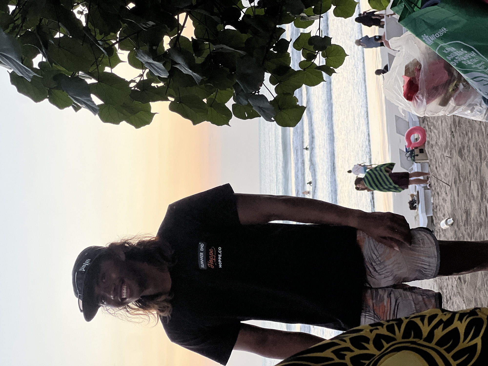 In Kuta, Indonesia, at sunset, a young man stands on the beach, facing left and smiling. He wears a black t-shirt with "SUMMER END HOPPE HOPPE.CO" printed on it, a black baseball cap, and patterned swim shorts. Behind him, the ocean is calm with a few surfers in the water. The beach is sandy, with lounge chairs, a pink float, and bags of trash scattered. Palm trees and other foliage frame the right side of the scene.