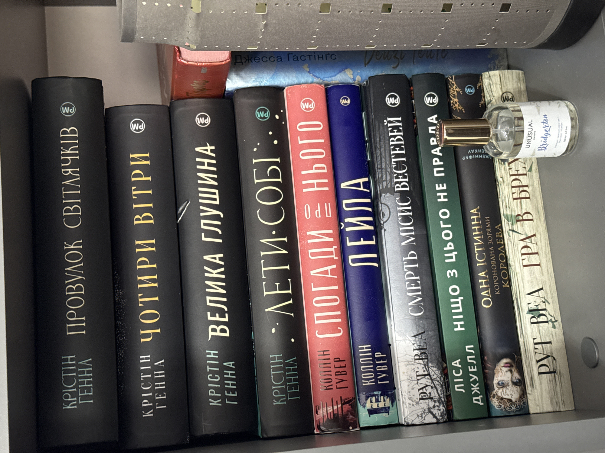 This is a high-angle, close-up photograph of a shelf filled with books and a small perfume bottle. The books are arranged vertically, with their spines facing the viewer. The spines are primarily black, with titles and author names in gold lettering. The titles appear to be in Cyrillic script. One book has a bright red cover, and another has a dark blue cover. A small, clear glass perfume bottle with a gold cap rests horizontally on top of two books towards the right side of the frame. The bottle has a label with text in English and what appears to be Polish. The background of the shelf is a muted gray, and a portion of what looks like a metal grate or decorative element is visible at the top of the image. The lighting suggests an indoor setting, likely a home or a store. The overall impression is one of organization and a personal collection of reading material.