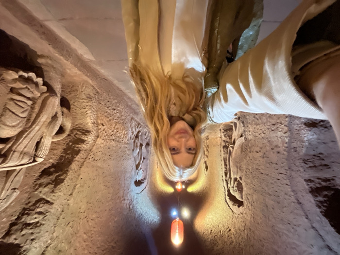 A selfie depicts a light-skinned female individual with long blonde hair, smiling directly at the camera. She wears a light-colored, long-sleeved top and a matching jacket, with her right arm extended, holding the camera. The scene is set within the Thousand-Buddha Cliff (Qianfo Grottoes) in Shizhong District, Guangyuan, Sichuan Province, China. The background features a rock-cut tunnel with extensively carved walls displaying numerous Buddhist figures and reliefs in various states of preservation. The textured, brownish-grey rock surfaces are illuminated by artificial light sources, including red lanterns and fixtures emitting warm orange and cool blue light, casting colorful glows on the carvings. A dark, level pathway is visible on the floor, extending into the illuminated passage. No other individuals are present.