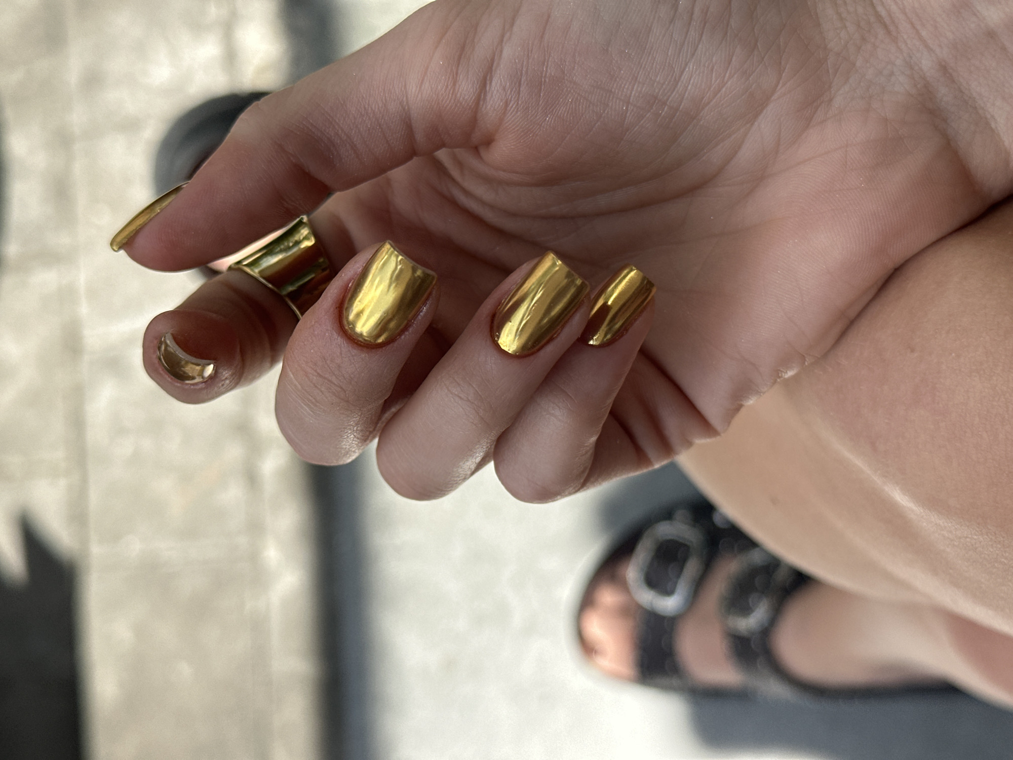 A close-up shot of a woman's hand with gold-painted nails and a large gold ring on her index finger. The woman is wearing black sandals with silver buckles. The background appears to be an outdoor paved surface. It is daytime. The city of Kerobokan Kelod, Indonesia, cannot be identified from the visual information.