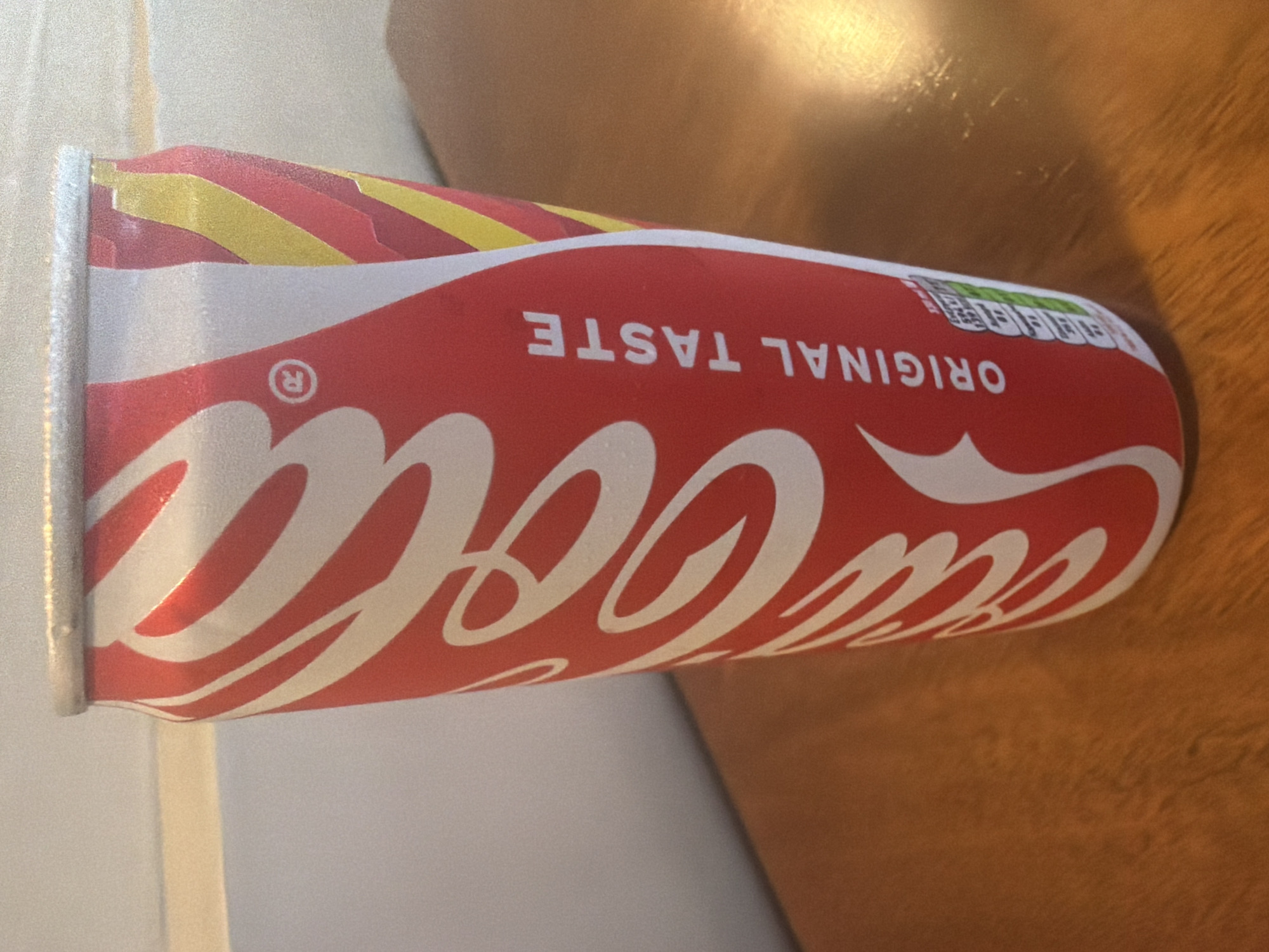 A close-up, low-angle shot shows a can of Coca-Cola with its iconic red and white branding. The can is positioned horizontally, with the "Coca-Cola" script prominently displayed in white against the red background. Above the script, the words "ORIGINAL TASTE" are visible in white letters. To the left of "ORIGINAL TASTE", a small registered trademark symbol "®" is visible within a white circle. The top of the can features diagonal stripes in red and yellow. In the upper right portion of the can, a rectangular nutritional information label is partially visible, with a green border and white text and icons. The background appears to be a wooden surface, possibly a table, with a light-colored surface to the left of the can. The lighting suggests an indoor setting, with a warm glow reflecting off the can and the wooden surface. The overall impression is a still life of a beverage product.