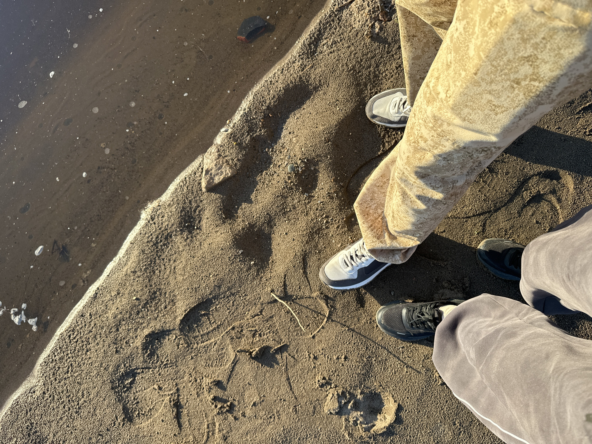 This is a high-angle, full-shot photograph of two individuals' legs and feet standing on a sandy shoreline next to a body of water. The sand is coarse and textured, with visible footprints. The water appears murky and still, with some small white frothy bubbles and darker debris visible on its surface. The individuals are wearing casual footwear: one has on dark-colored sneakers and grey sweatpants, while the other is wearing light grey and dark blue sneakers with patterned tan pants. The sunlight casts long shadows across the sand, indicating it is either early morning or late afternoon. The scene appears to be located on the shore of a river or lake in Noviy, Russia.