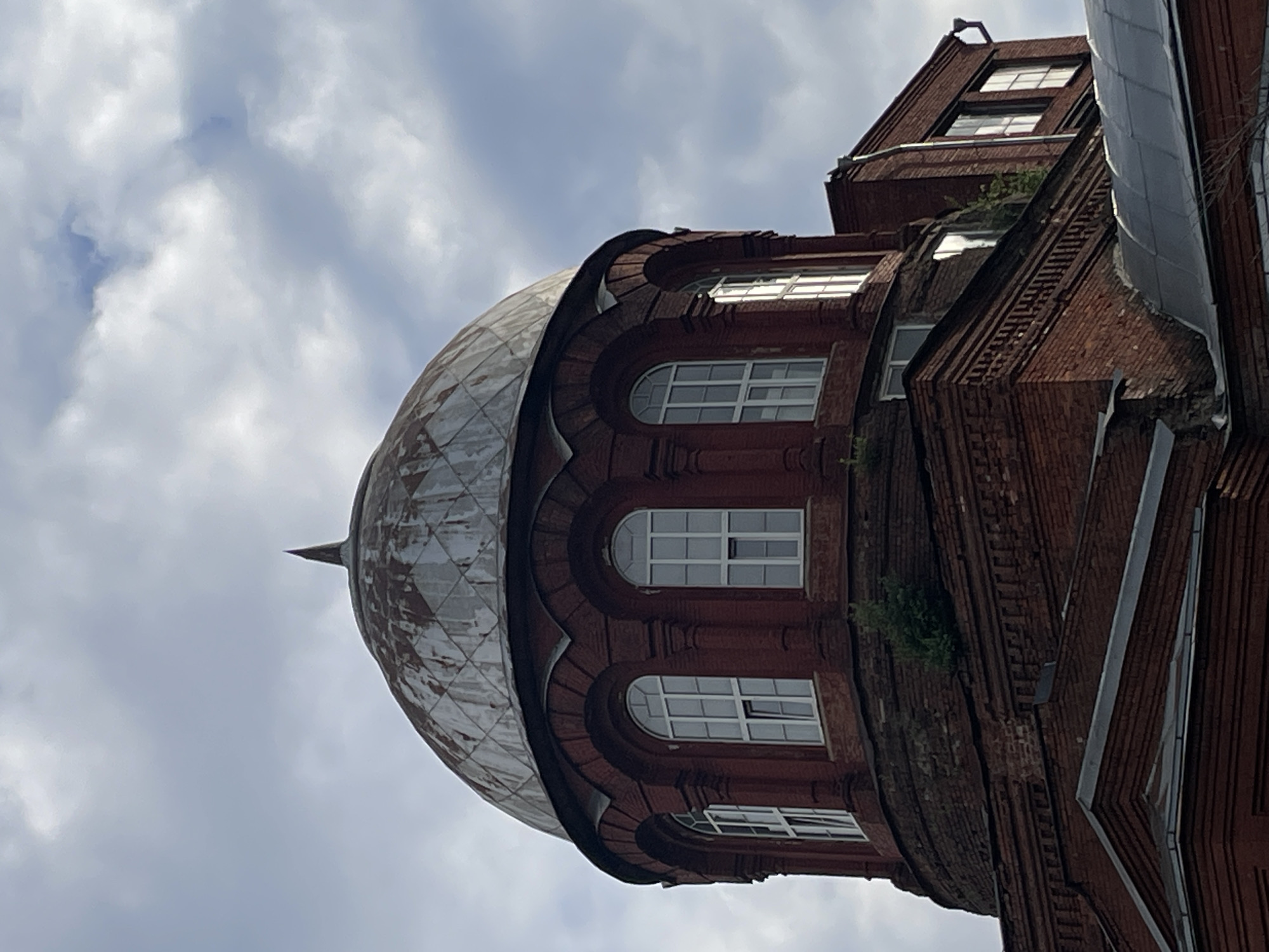 The image shows the dome and upper levels of a red brick building with a weathered, diamond-patterned metal roof topped by a pointed spire. The dome is supported by a cylindrical base with arched windows, each divided into multiple panes by white frames. The brickwork is a deep red, and some areas show signs of age and wear. Green foliage is visible growing from crevices in the brickwork on the lower sections of the dome and adjacent building sections. To the right of the dome, a rectangular brick structure with similar arched windows is attached. The sky above is cloudy with patches of blue. Based on the architectural style, the building appears to be a church or a similar religious or historical structure. A quick search indicates this is the Holy Trinity Church in Sestroretsk, a suburban town in the north-western part of Saint Petersburg, Russia, not Moscow.