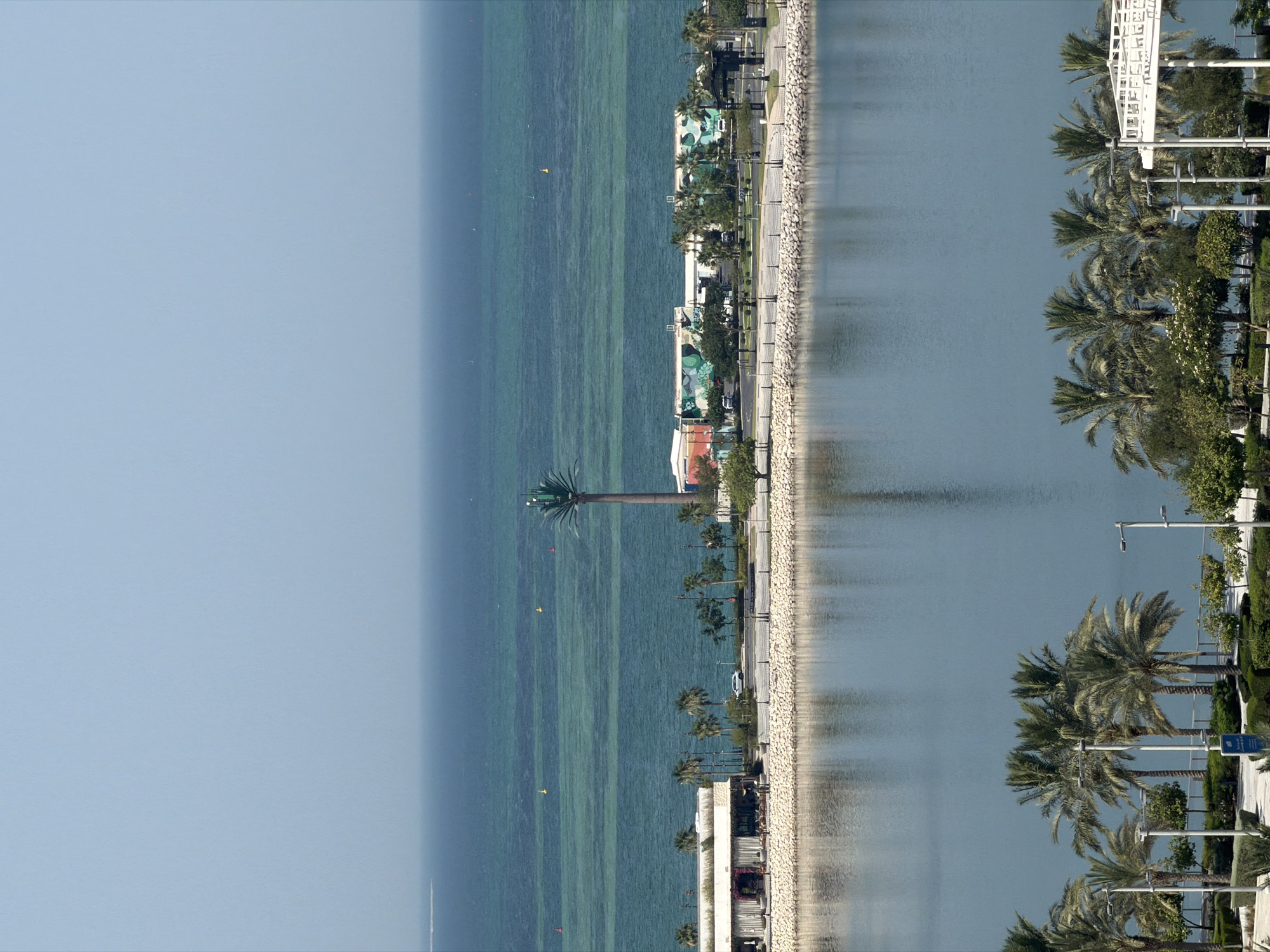 An elevated, high-angle, full shot captures a coastal scene with a body of water in the foreground and the sea in the background, under a clear, pale blue sky. The foreground is dominated by a calm, reflective body of water, with the faint reflections of trees and structures visible on its surface. Beyond this, a paved walkway lines the edge of the water, bordered by a low stone wall and an array of palm trees and manicured bushes.
Further back, a row of buildings with white, modern architecture and flat roofs stretches along the coastline. Some of these buildings feature large, colorful murals on their sides. A single white SUV is parked on the paved area between the buildings and the water's edge. Beyond the buildings, the turquoise sea stretches to the horizon. Several buoys are visible in the shallow, clear water near the shore, indicating a protected area or a marker. A tall, slender structure resembling a palm tree with a communications array at its top stands prominently on the coastline, between the buildings and the sea. The overall impression is one of a developed, resort-like coastal area.