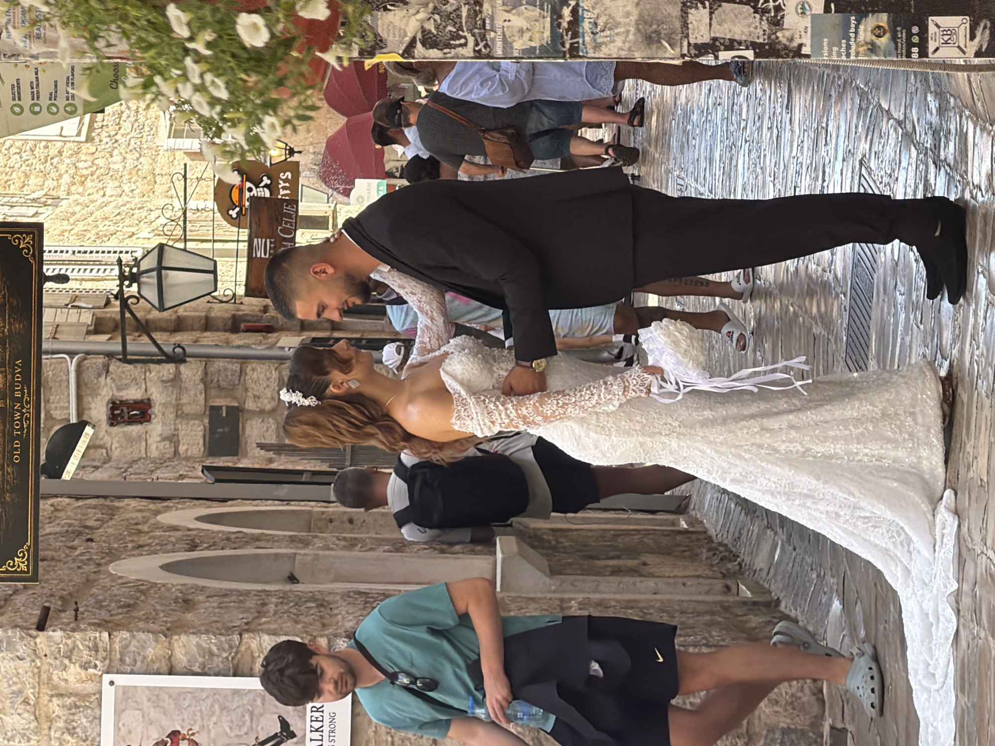 The image captures a wedding scene in the old town of Budva, Montenegro. A newly married couple, the bride in a white lace gown and the groom in a black suit, stand facing each other on a narrow, cobblestone street lined with stone buildings. The bride's dress features long, fitted lace sleeves and a full skirt that trails onto the wet cobblestones. She wears a floral hairpiece. The groom is embracing her, his left arm around her waist.
In the background, several other people are visible, some walking and others standing. To the left, a young man in a teal t-shirt and black shorts holds a bottle of water. Behind him, another man with a backpack walks away from the camera. Further down the street, other individuals in casual attire can be seen, some holding red umbrellas. The environment suggests a warm, sunny day. Above the couple, signs are visible, including one that reads "OLD TOWN BUDVA" and another with a skull and crossbones graphic. The cobblestone street appears wet, reflecting the surrounding light.