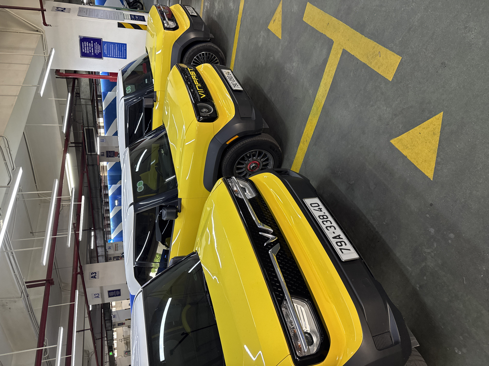 Three yellow VinFast electric cars are parked in a row in an indoor parking garage. The cars are facing towards the viewer, and are positioned diagonally within the parking spots, indicated by yellow painted lines on the concrete floor. The license plates of the cars are visible, with the closest car displaying "79A-338.40", and the second car showing "79C-090.84". In the background, there are structural beams and lighting fixtures overhead, and signs indicating parking sections labeled "A3" and "B4". The environment appears to be a well-lit, modern parking facility, potentially in Nha Trang, Vietnam, given the license plate format. There are no people clearly visible in the image.