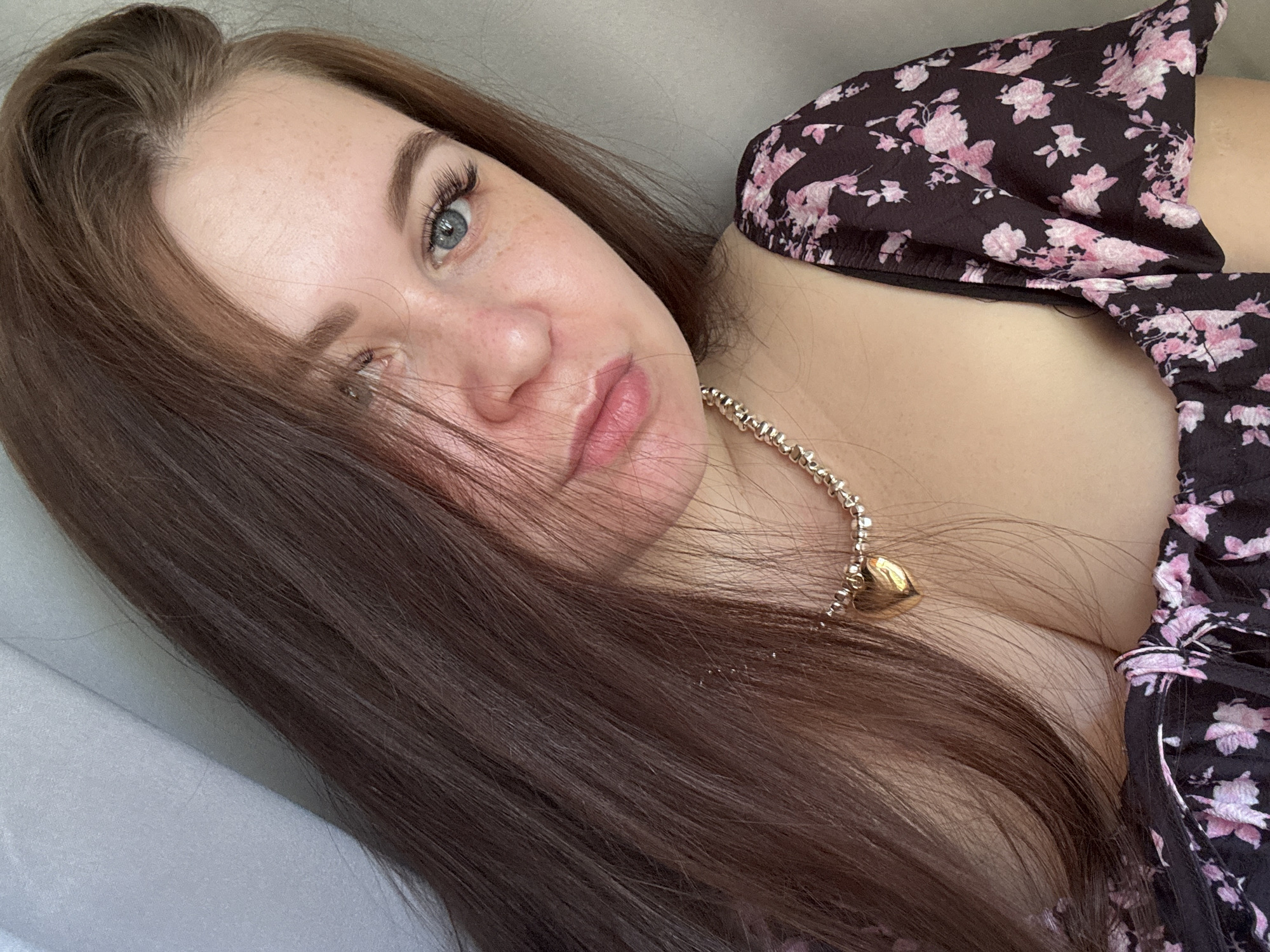 The image is a close-up portrait of a young woman with long, brown hair and blue eyes. She is wearing a dark floral-print top and a gold necklace with a heart-shaped pendant. Her skin has light freckles. The background is a neutral, light gray surface. There is no indication of location in Chaikovsky, Russia, within the image itself.