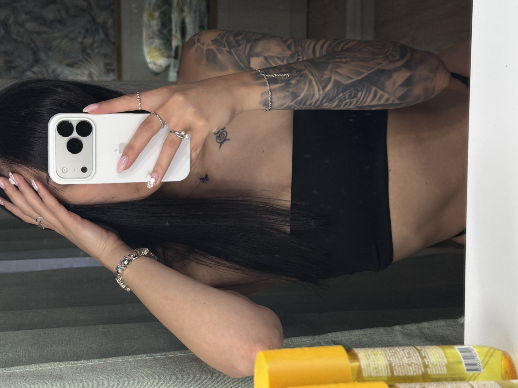 A young adult female is depicted taking a mirror selfie indoors in Plovdiv, Bulgaria. She has long, dark hair, partially obscuring her face, and light-toned skin. She is wearing a black strapless top. Her left arm features extensive gray-wash tattoos, covering the entire sleeve from shoulder to forearm with intricate designs. Smaller black tattoos, including a butterfly and an abstract symbol, are visible on her chest. Her nails are manicured with light pink polish and white floral accents.
She holds a white smartphone, likely an iPhone in a white case, with her right hand, positioned to capture her reflection. Her right hand displays multiple silver rings and a silver chain bracelet with various charms. Her left hand is raised to her head, with a silver ring on her finger and a silver bracelet visible on her wrist. A delicate silver chain necklace with a small dark blue butterfly pendant adorns her neck.
In the foreground, two yellow plastic bottles with partial labels and a barcode are visible on a white surface, which serves as a counter or shelf. The background of the reflection reveals a dark green curtain to the left and a wall with a muted, patterned texture, possibly wallpaper or artwork, to the right. Further right in the background, a light-colored textile, possibly bedding, is visible on a surface. The setting appears to be an interior room, likely a bedroom or dressing area.