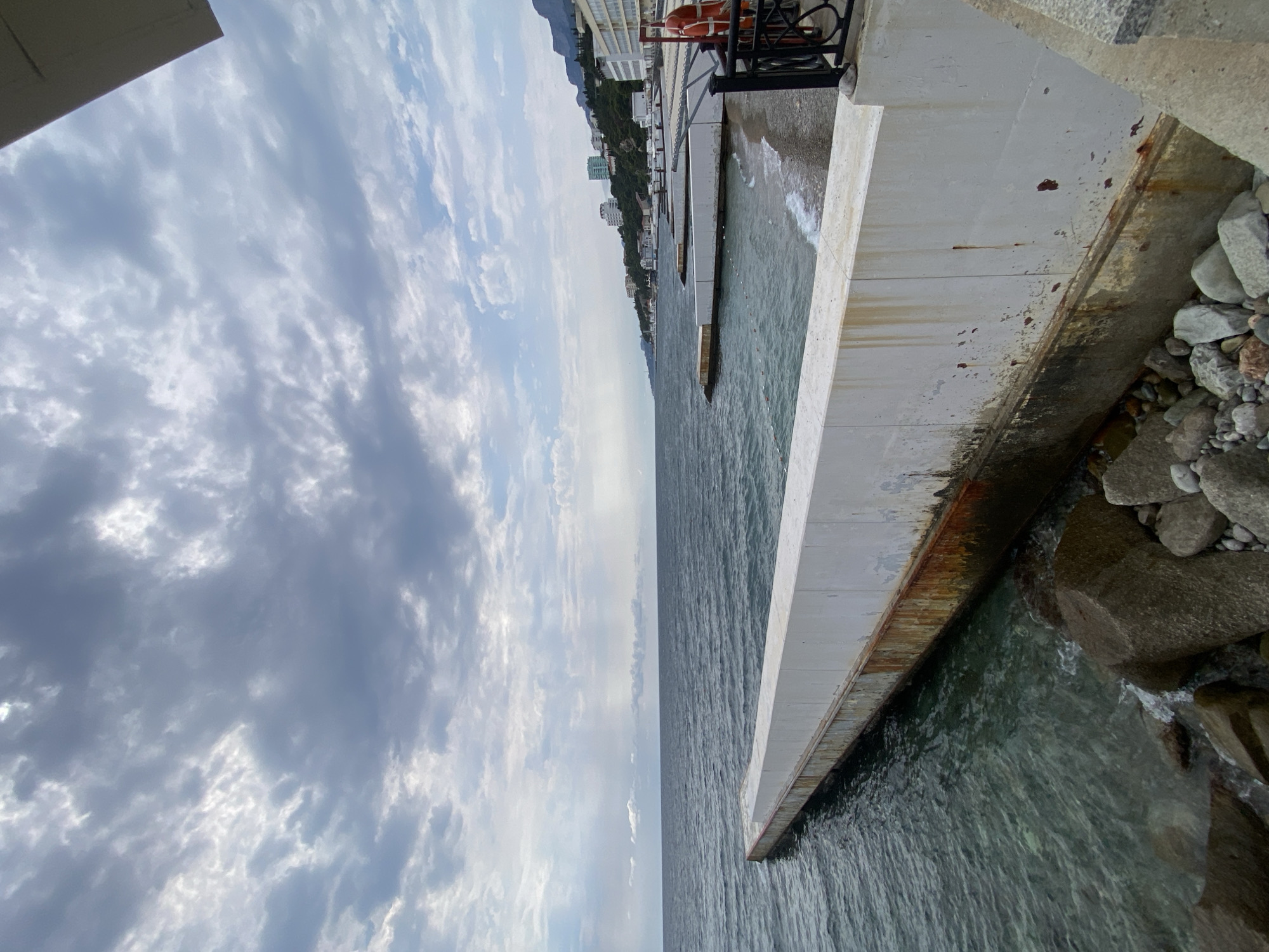 The image shows a concrete pier extending into a body of water under a cloudy sky. The pier is weathered, with visible rust stains and damage to its surface. Large rocks are scattered at the base of the pier. In the distance, a coastline is visible with buildings and a mountainous backdrop. The water is a deep blue-green color. The scene suggests a coastal area, likely a resort or waterfront promenade. The provided information states the location is in the village council of Gaspra, Ukraine.