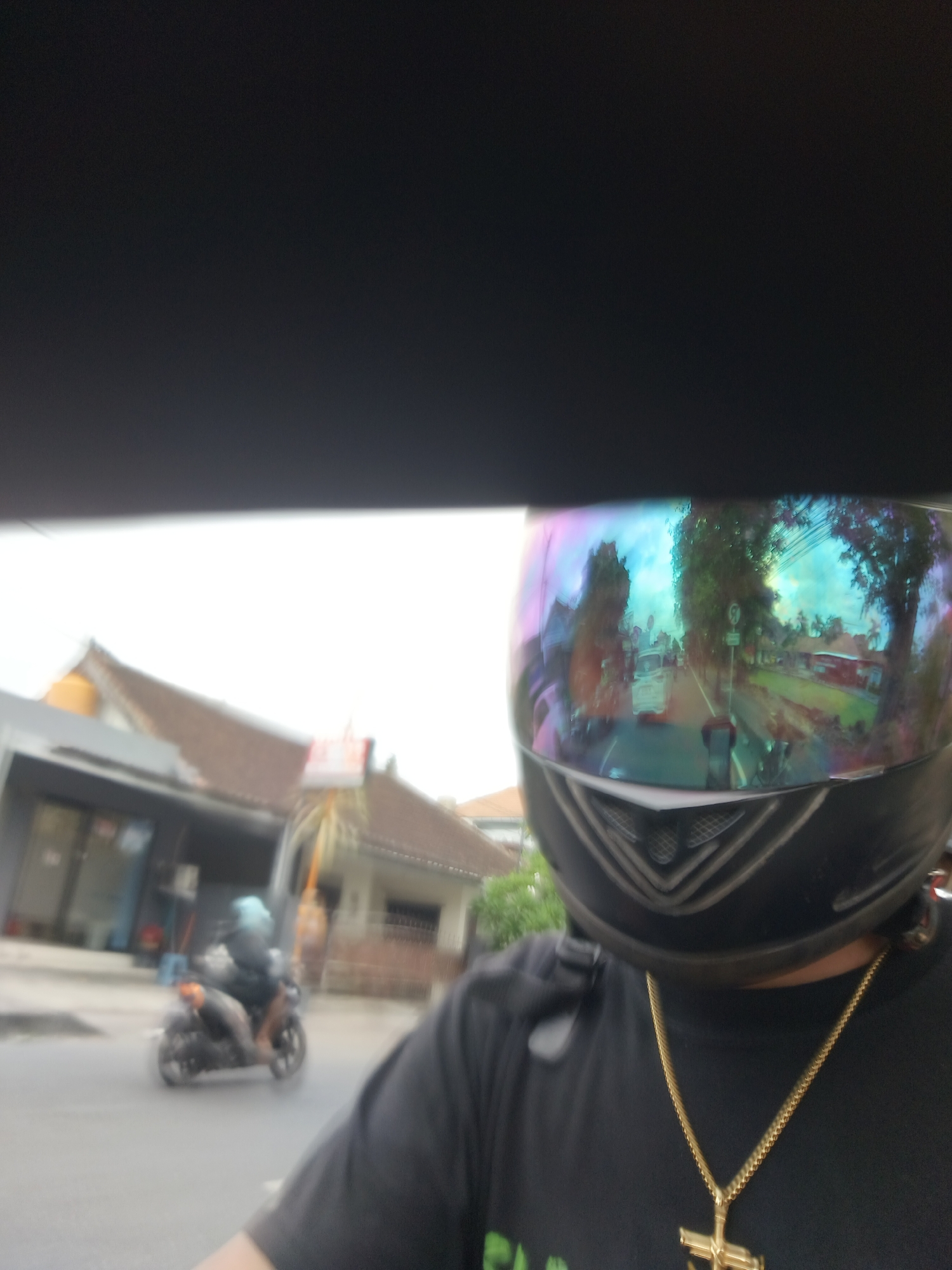 A first-person perspective selfie shows a person wearing a black t-shirt and a motorcycle helmet with a reflective visor. The visor reflects a street scene with vehicles, trees, and buildings, suggesting an urban environment, likely Denpasar, Indonesia, given the architectural style of the buildings and the presence of a scooter on the road. The person is wearing a gold chain with a cross pendant. In the background, a scooter with a rider is visible on the road. The image has a slight blur, common in action shots or from a moving vehicle.
