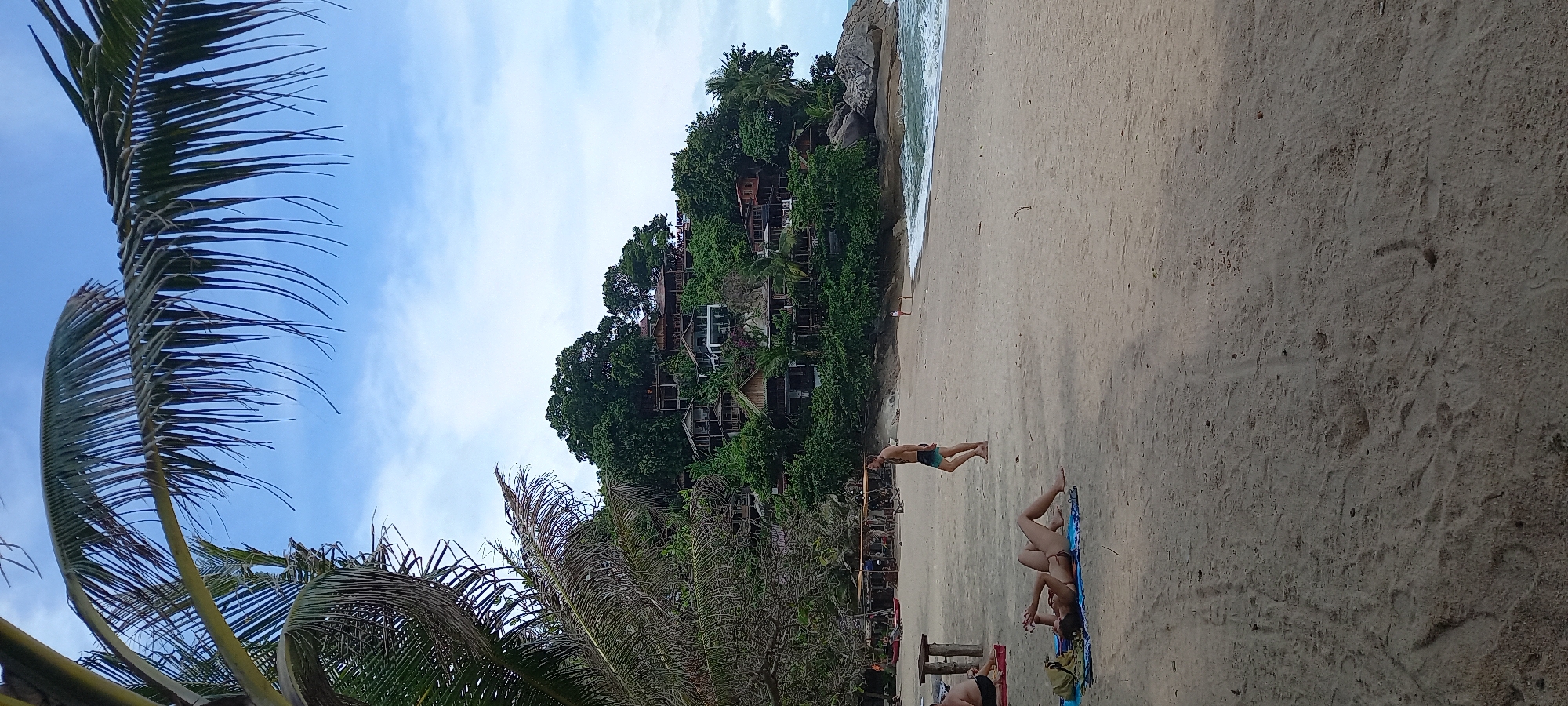 The image shows a beach scene in Baan Thong Nang, Thailand. The beach has light-colored sand and slopes up to lush vegetation and some buildings on a small hill. A man is walking near the shoreline. In the foreground, a woman is lying on a blue towel on the beach. A group of people are sitting by some red and black objects nearby. Palm trees are visible on the left side of the frame, partially obscuring the view. The sky is light blue with some clouds.