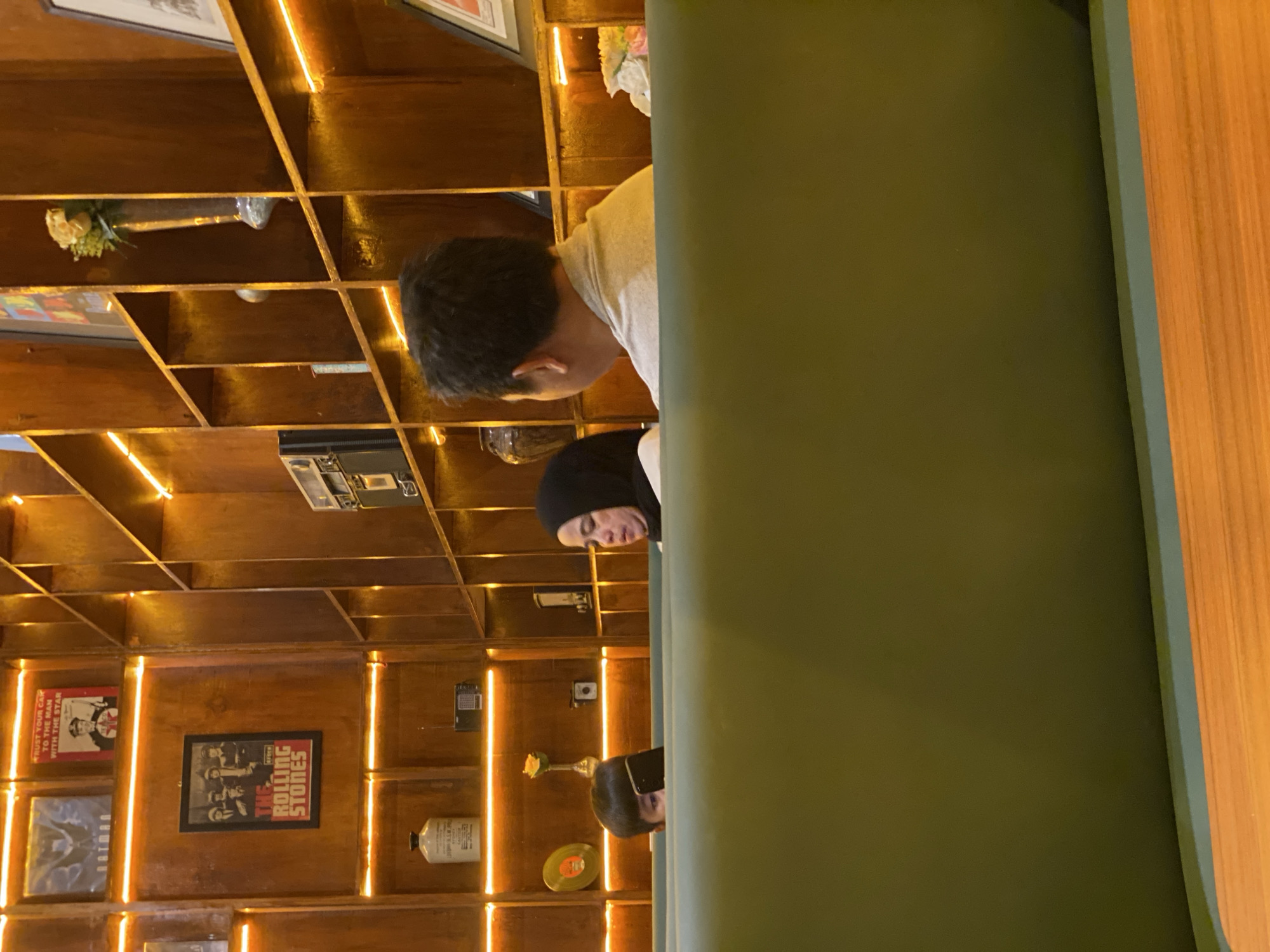 The image shows an indoor scene, likely a cafe or restaurant, with wooden shelves and warm lighting. In the foreground, a person with dark hair is partially visible, looking at a black smartphone. To their right, a woman wearing a black hijab is looking to the side. Behind them, a man in a light-colored shirt is facing away from the camera. The shelves are decorated with various items including framed pictures, what appears to be a record, and a vase with a flower. One framed picture displays the text "THE ROLLING STONES." The environment appears to be dimly lit with yellow LED strip lights illuminating the shelves. The photo is taken from a low angle, emphasizing the seating area and the shelves. The setting is in the city of Banjarbaru, Indonesia, suggesting a public establishment.