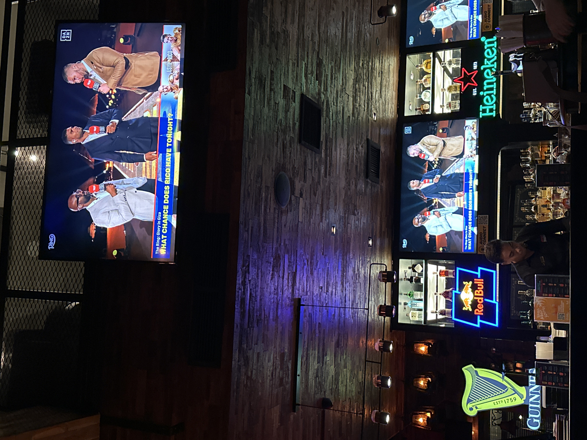 The image shows the interior of a bar or entertainment venue with multiple televisions displaying a boxing match. Several neon signs are visible, advertising Heineken, Red Bull, and Guinness. One person is seen behind the bar, presumably a bartender. The walls are decorated with a wood panel design. The image suggests a lively, pub-like atmosphere. The image does not provide information about North Kuta, Indonesia.