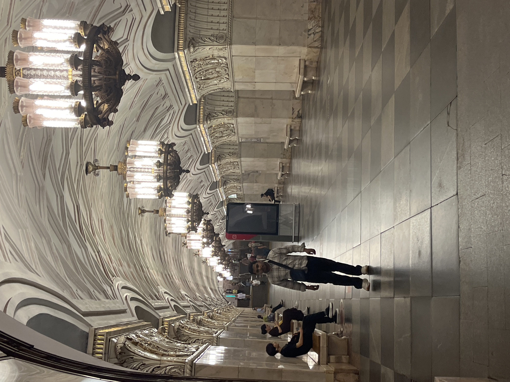 The image depicts the interior of a Moscow Metro station, likely a grand one, characterized by its ornate architecture. The station features a high, arched ceiling adorned with intricate patterns and decorative moldings in shades of white, grey, and gold. Large, elaborate chandeliers with multiple glass tubes hang from the ceiling, illuminating the space. The walls are lined with polished marble columns featuring detailed gold ornamentation. Along the walls, there are benches for passengers. The floor is paved with large, checkered tiles in shades of grey and dark grey. Several people are visible in the scene. In the foreground, a man in a plaid shirt and dark pants is walking towards the camera. Further down the platform, other individuals are seated on benches or walking, including a few people on stairs leading to another level. A large digital display screen is mounted on a column. The overall impression is one of opulence and grandeur, typical of many historic Moscow Metro stations.