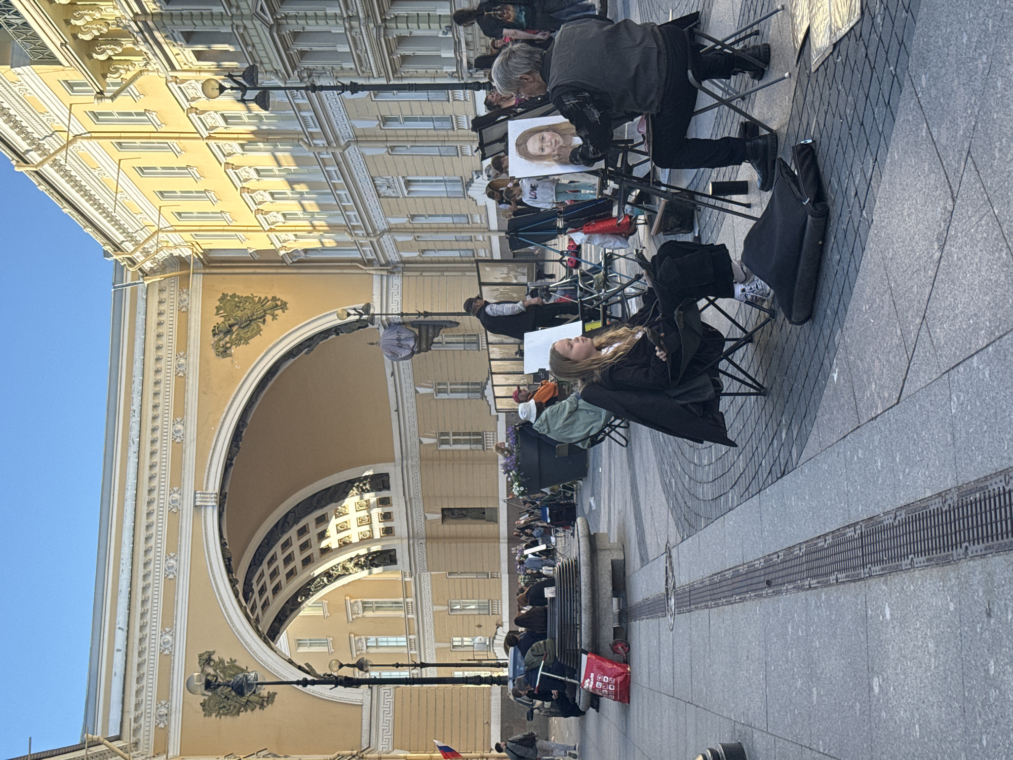 The image depicts a sunny day in Palace Square, Saint Petersburg, Russia, with the General Staff Building's triumphal arch dominating the background. In the foreground, an artist, an elderly man with graying hair, is seated, painting a portrait of a young girl. The girl, wearing a black jacket and white sneakers, sits patiently in a folding chair, looking towards the artist. Several other easels, art supplies, and folding chairs are set up in the open square, suggesting an area where artists typically work. In the middle ground, a group of people are seated on a bench, some looking towards the artists, while others are engaged in conversation. A red bag with a trolley and a scooter are visible near the bench. The architecture of the surrounding buildings is classical, characterized by yellow facades, white detailing, and numerous windows. The clear blue sky and the long shadows cast by the buildings indicate it is late afternoon. The overall atmosphere is one of a public gathering with artistic activity.